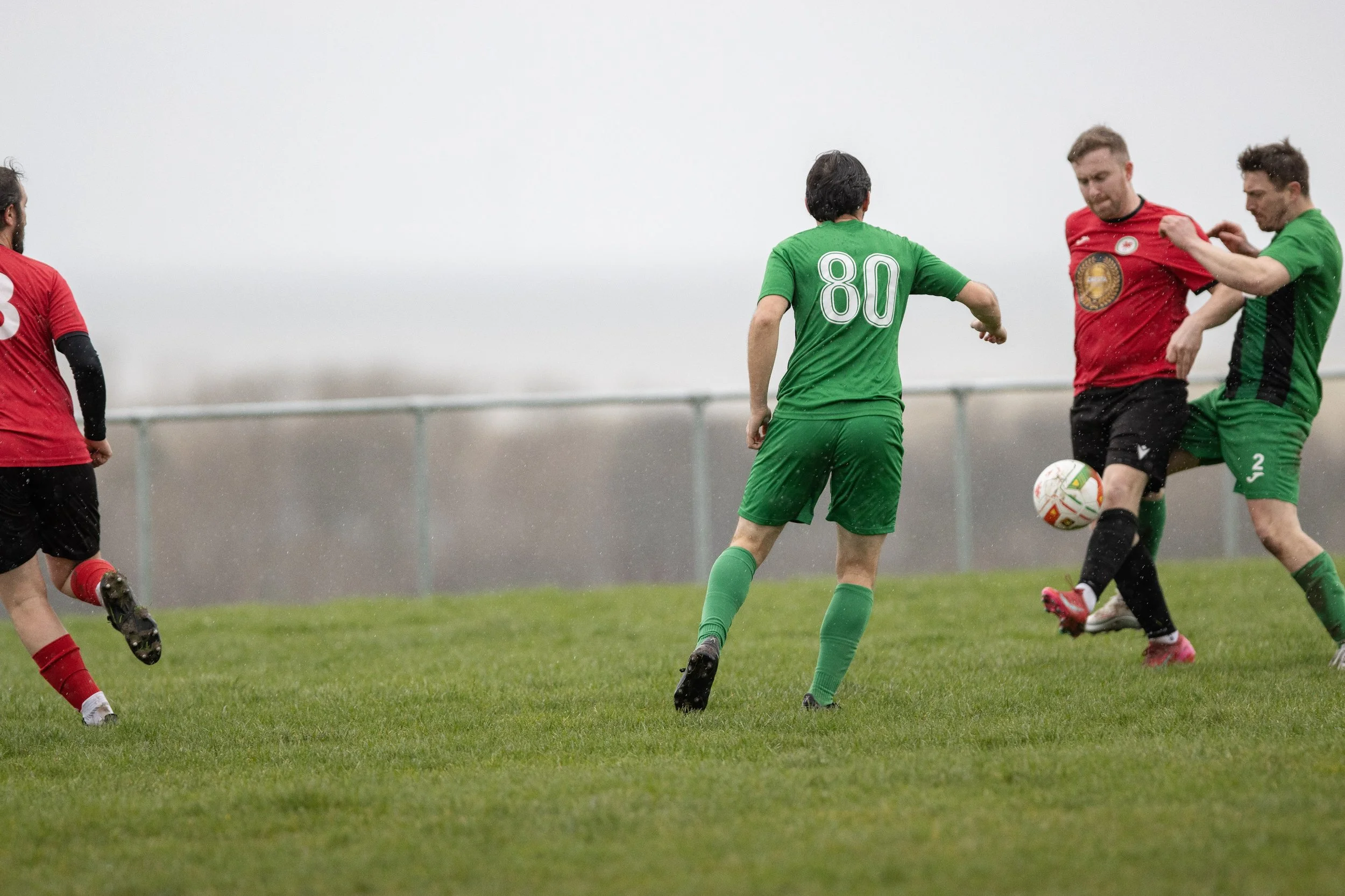 Four men playing soccer on a grassy field on a rainy day, two wearing green jerseys and two wearing red jerseys, with one man in green numbered 80 and another in green numbered 52, all pursuing the ball.