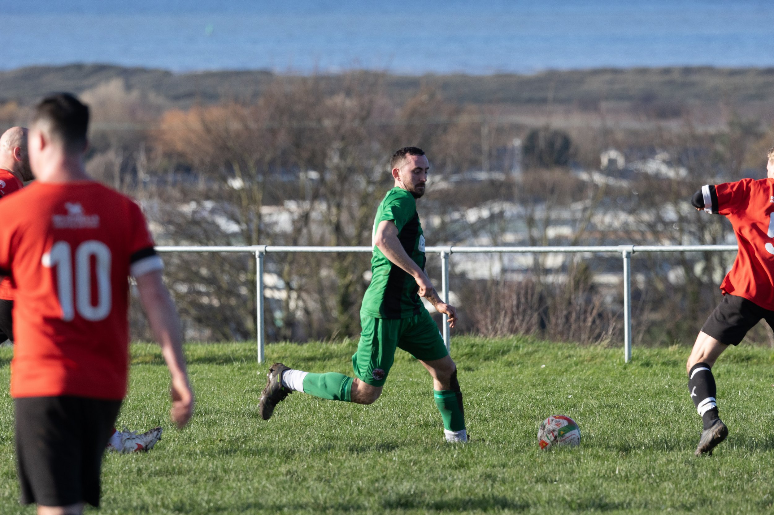 Soccer players on a grassy field during a game, with a man in a green jersey and black shorts in the center, running towards a soccer ball, while other players in red jerseys are nearby, and a metal railing, trees, and a distant hillside are visible 
