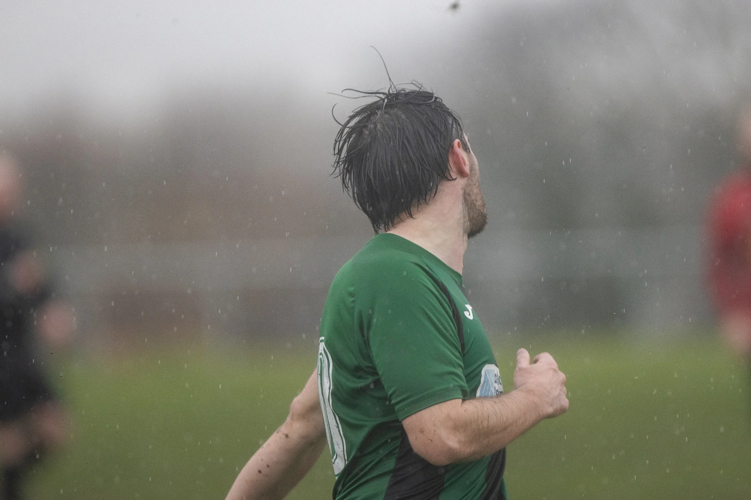 A male soccer player with dark, wet hair, wearing a green jersey, running in the rain during a match.