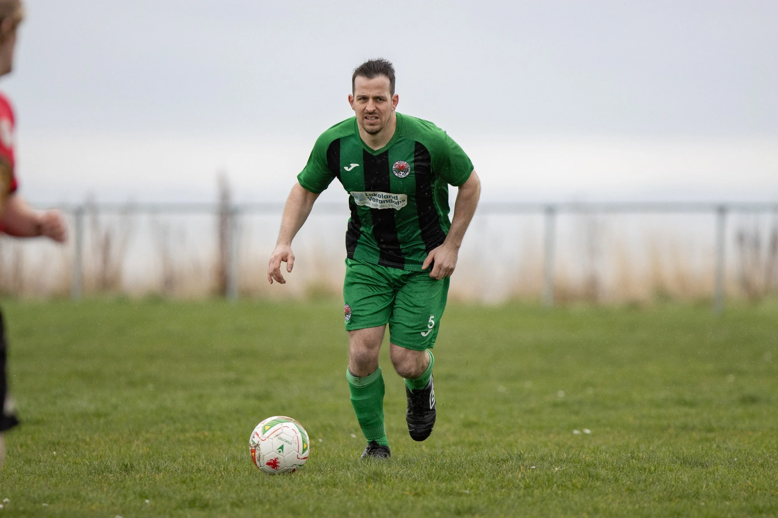A male soccer player wearing a green and black uniform with the number 5 on his shorts, running on a grass field with a soccer ball at his feet. The background features a cloudy sky and a blurred fence.