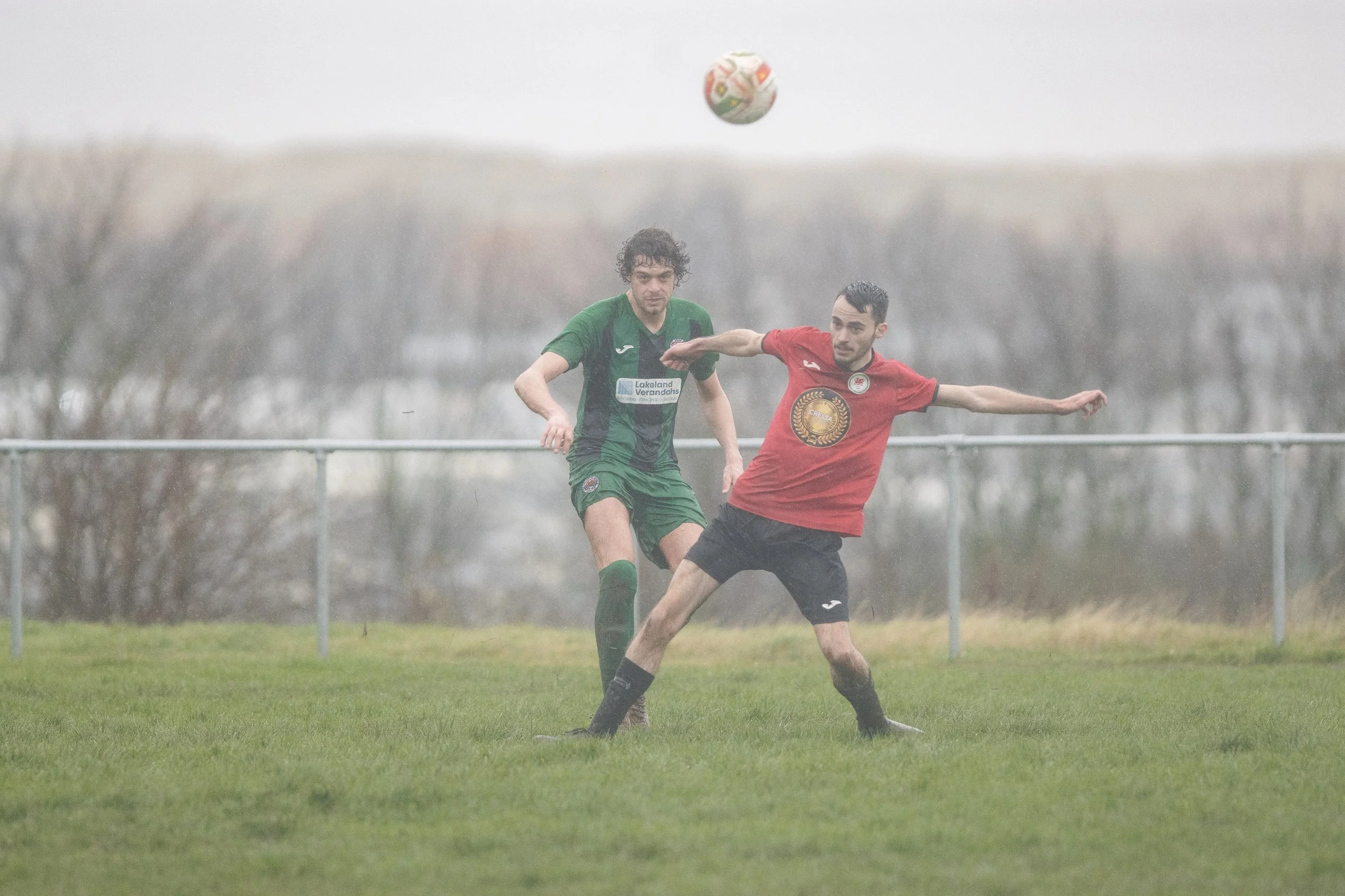 Two soccer players in green and red jerseys compete for the ball on a rainy field with blurred trees in the background.