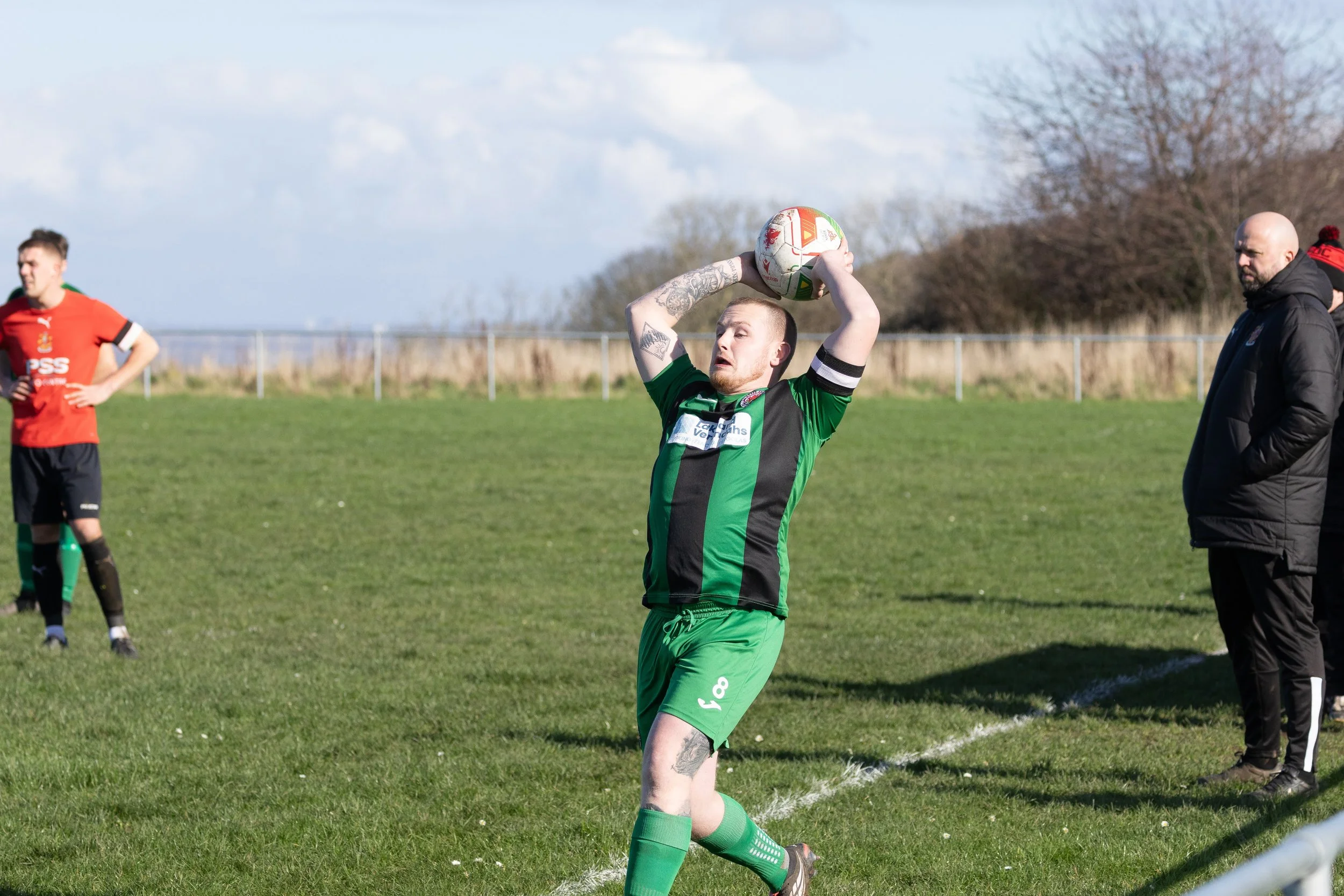 A soccer player in a green and black uniform is preparing to throw the ball back into play during a game on a grassy field. Two other players and a coach or referee are visible in the background, along with a fence and trees.