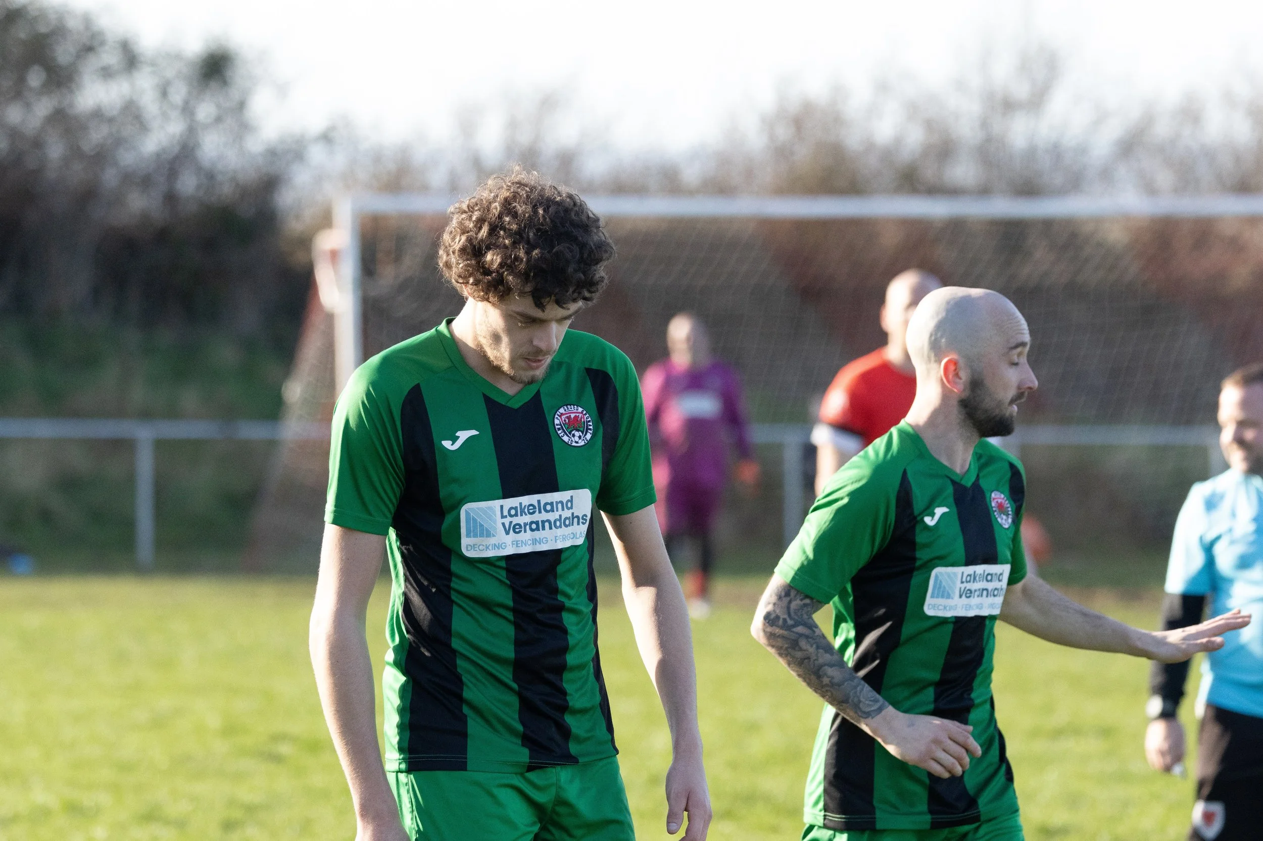 Two male soccer players in green and black uniforms walking on a soccer field, with a soccer goal and other players in the background.