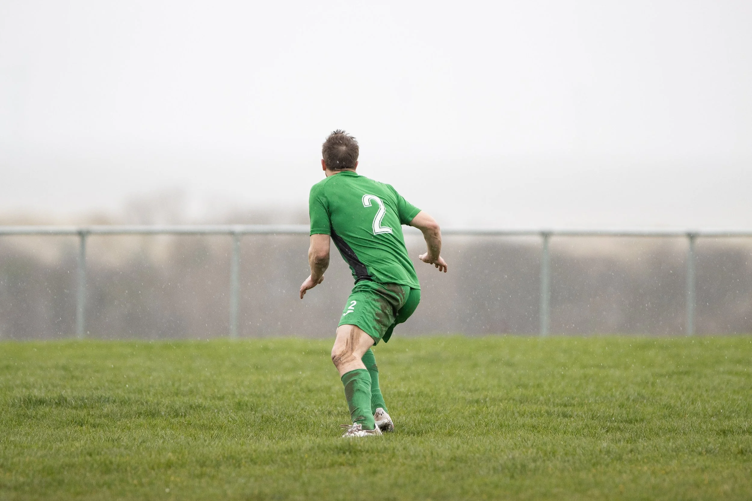 A soccer player wearing a green uniform with the number 2 on the back, running on a grass field, with a blurred railing and gray sky in the background.