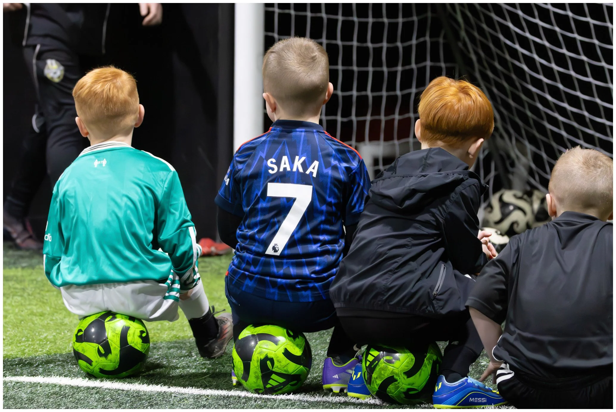 Four young children sitting on their knees on a soccer field while watching a game, with soccer balls under their knees. They are wearing sports clothing, including jerseys and jackets, with one child's jersey displaying the name "SAKA" and the numbe