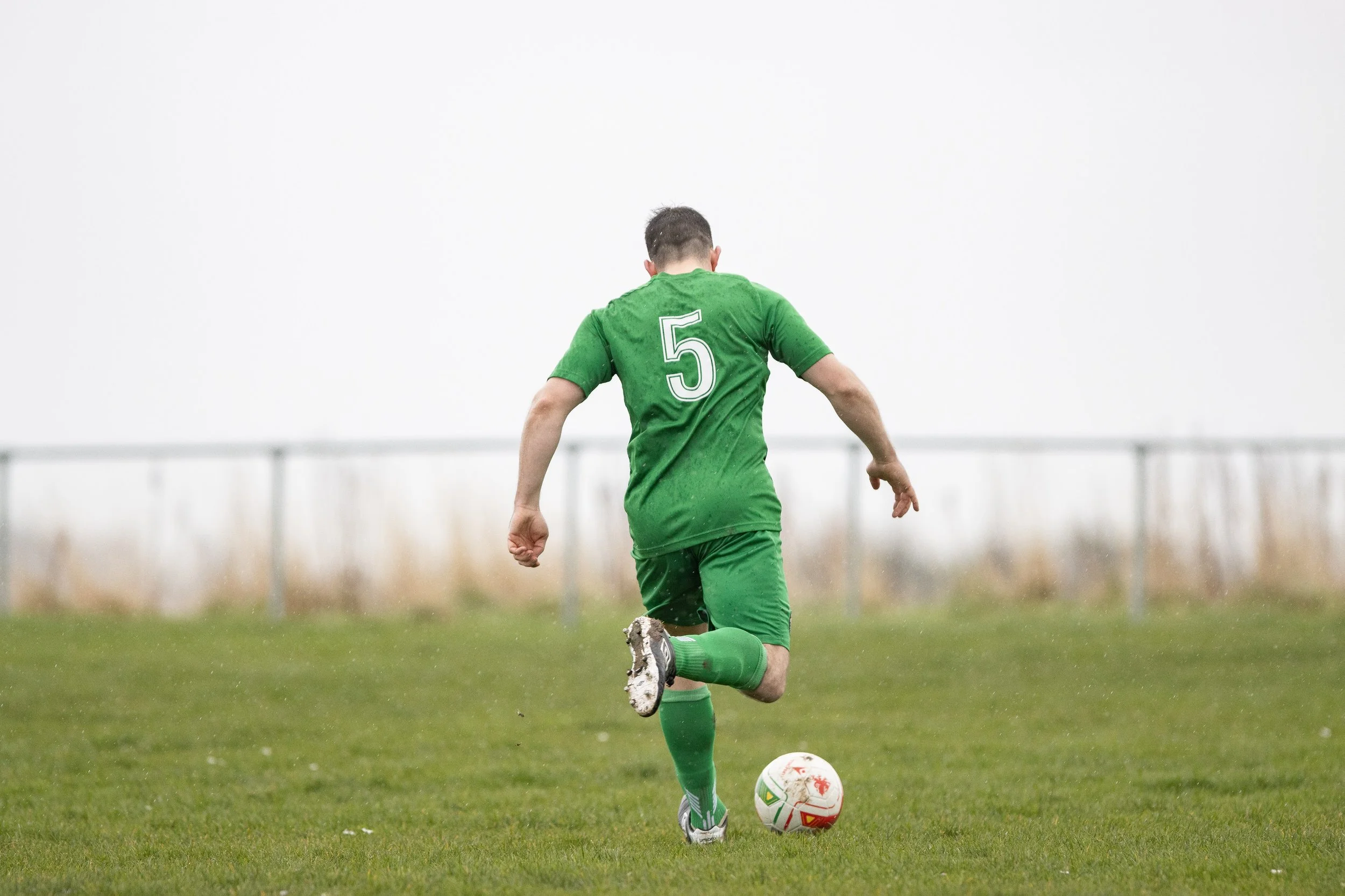 A soccer player in a green uniform, with the number 5 on the back, kicking a soccer ball on a grassy field.