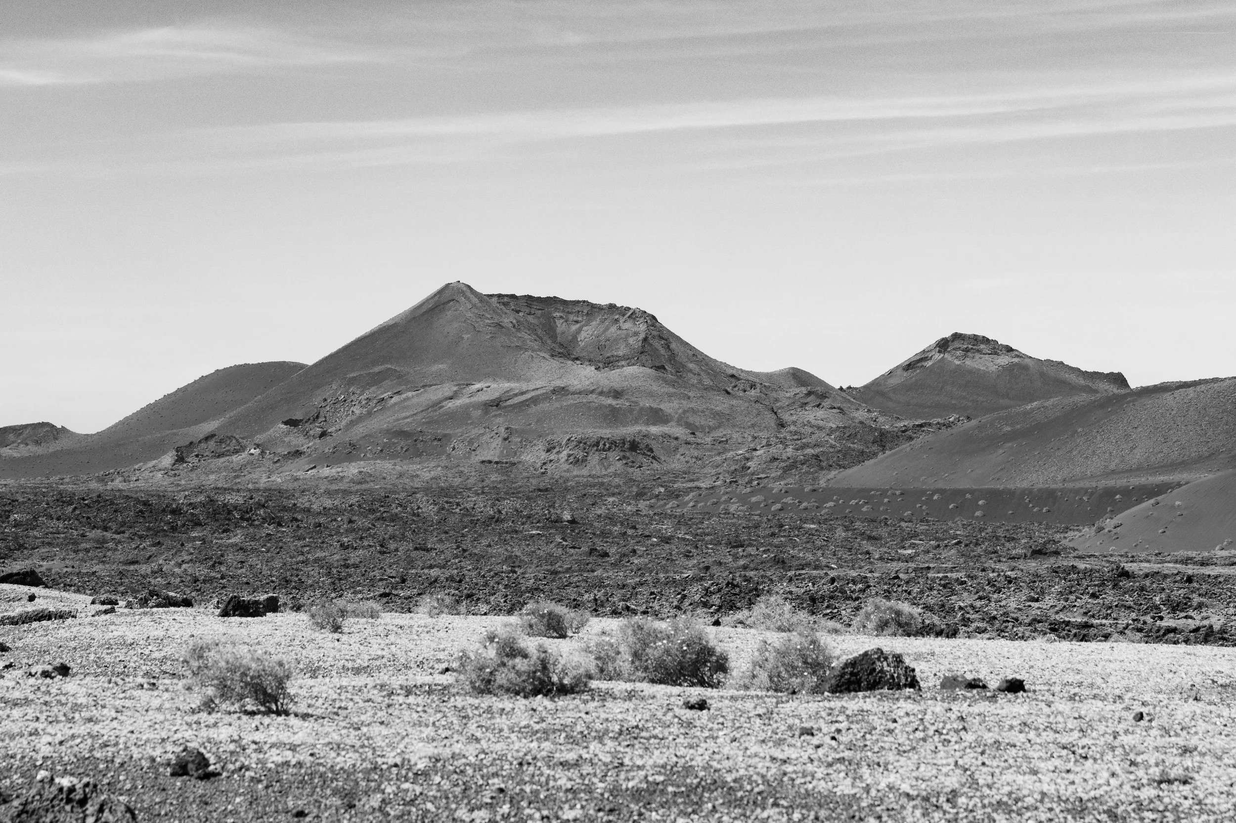 A black and white photo of a desert landscape with mountains in the background, scattered shrubs, rocks, and uneven terrain under a partly cloudy sky.