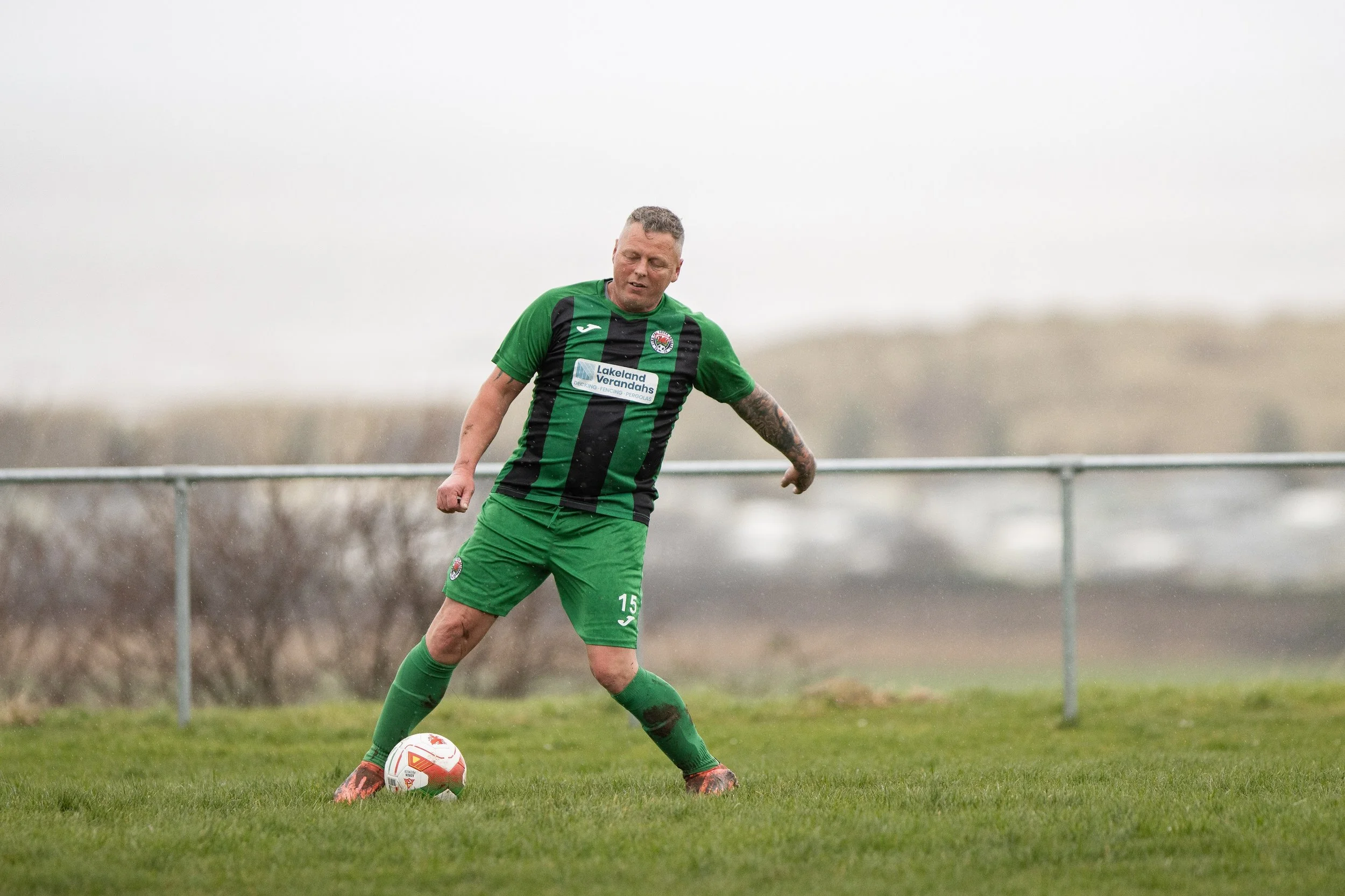 Male soccer player in green and black uniform kicking a soccer ball on a grassy field with a fence in the background.