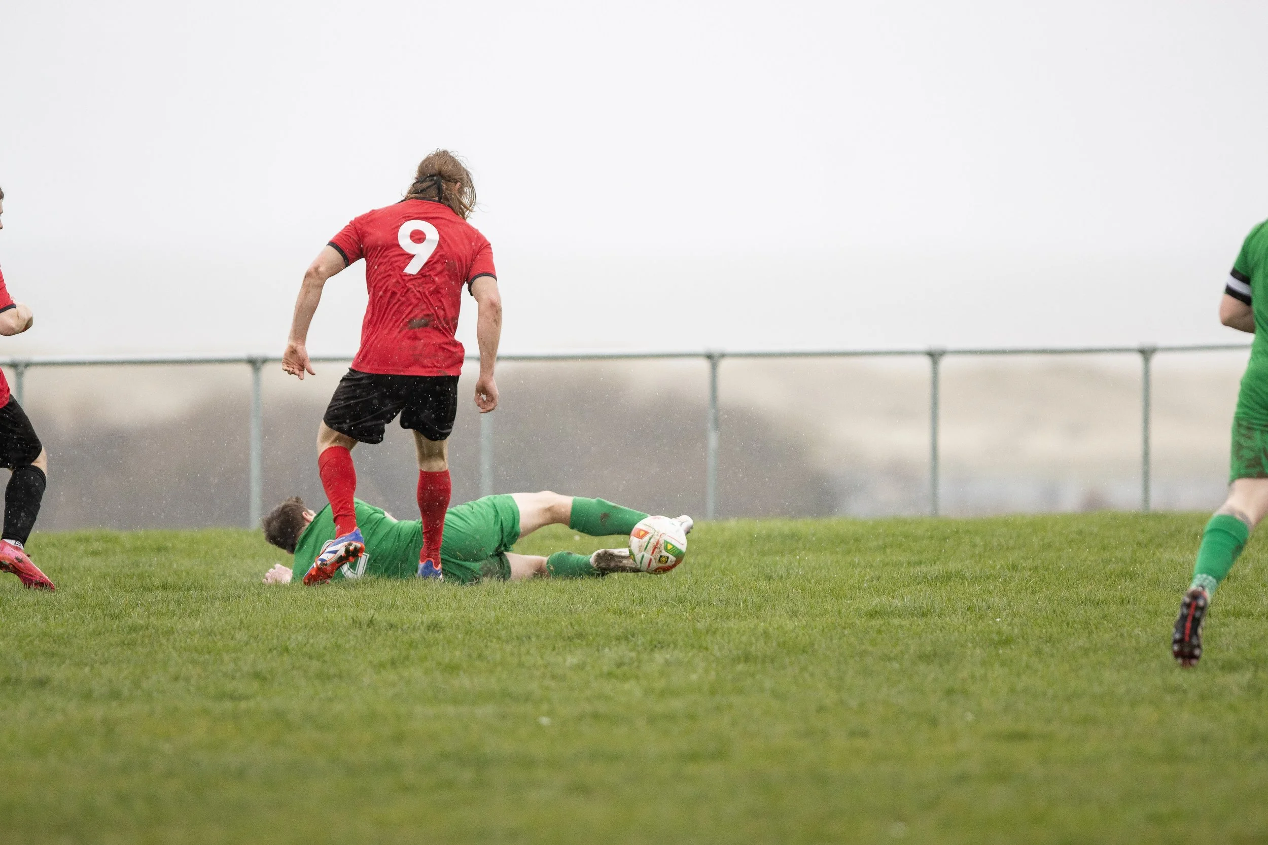 Soccer players in action on a grassy field with one player in green lying on the ground and others standing nearby, in rainy weather.