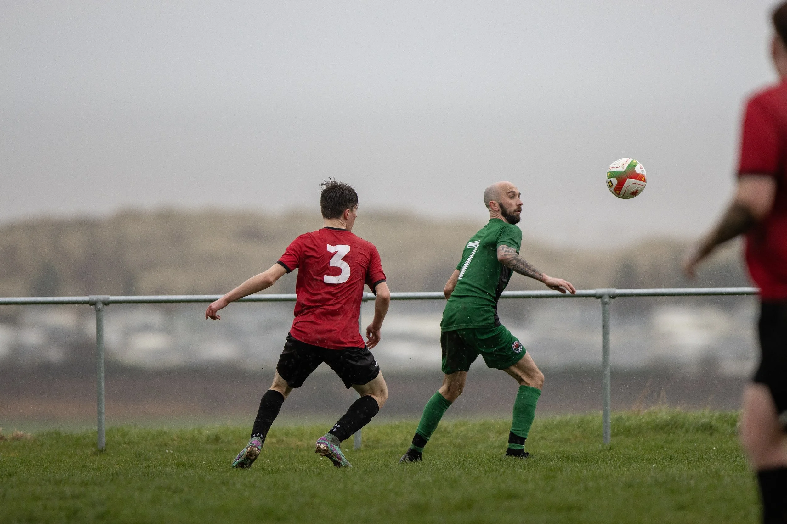 Soccer players in red and green jerseys playing on a grassy field in rainy weather, with one player heading the ball.