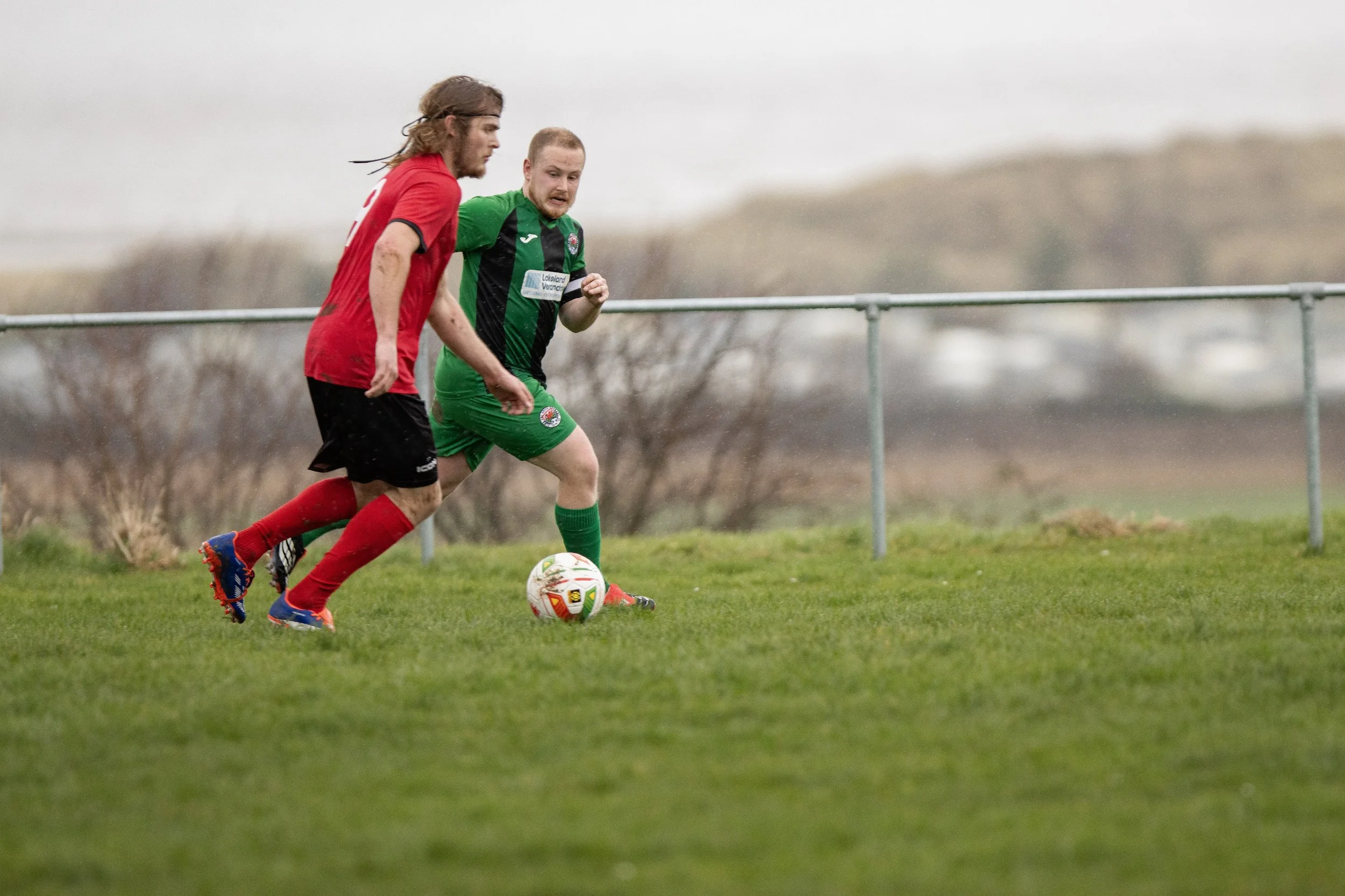 Two soccer players are competing for the ball on a grassy field, one in a red jersey and black shorts, and the other in a green and black jersey with green shorts.