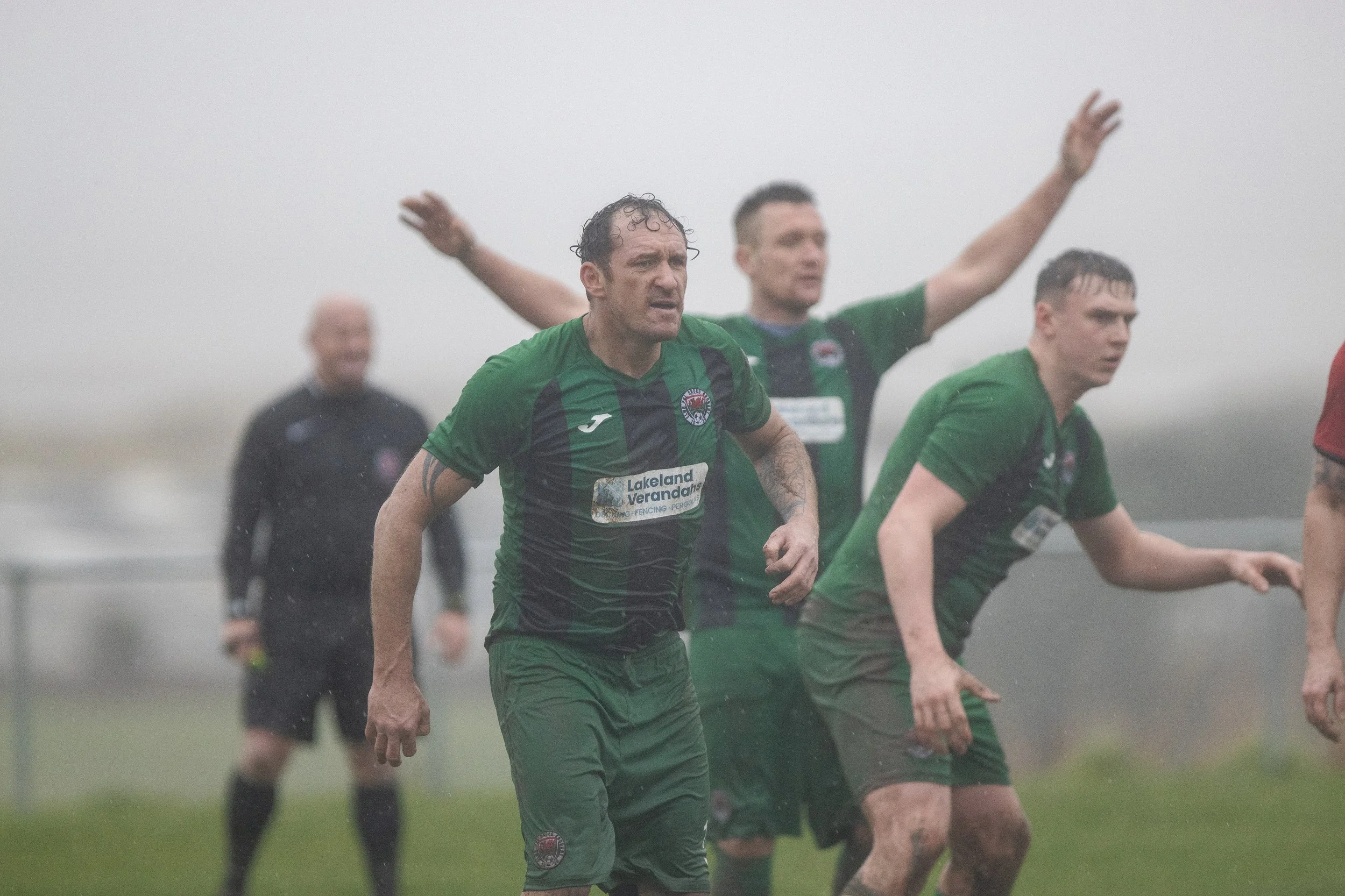 Soccer players in green uniforms running in pouring rain, with one player in the forefront, and a referee in black in the background.