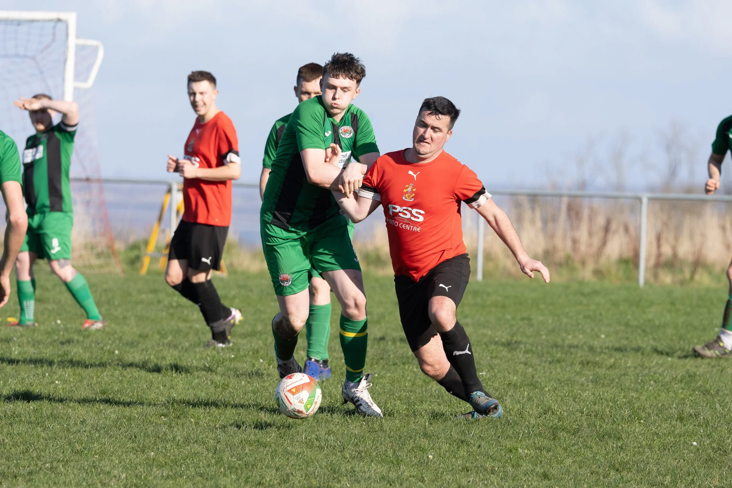 Two soccer players, one in green and one in red, compete for the ball on a grassy field with other players in the background.
