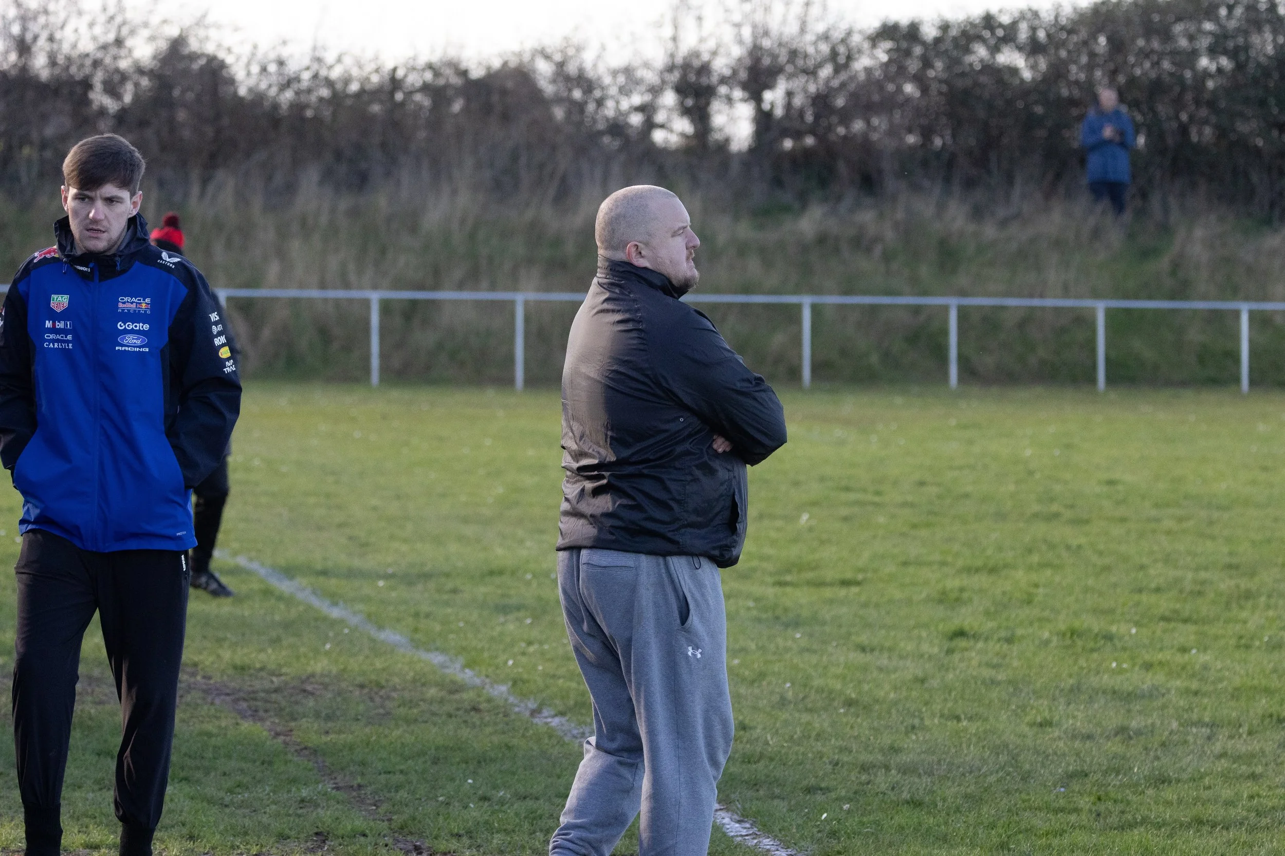A man with a shaved head wearing a black jacket and gray sweatpants stands with arms crossed on a grassy sports field, with another young man in a blue jacket nearby, and a woman in the background observing.