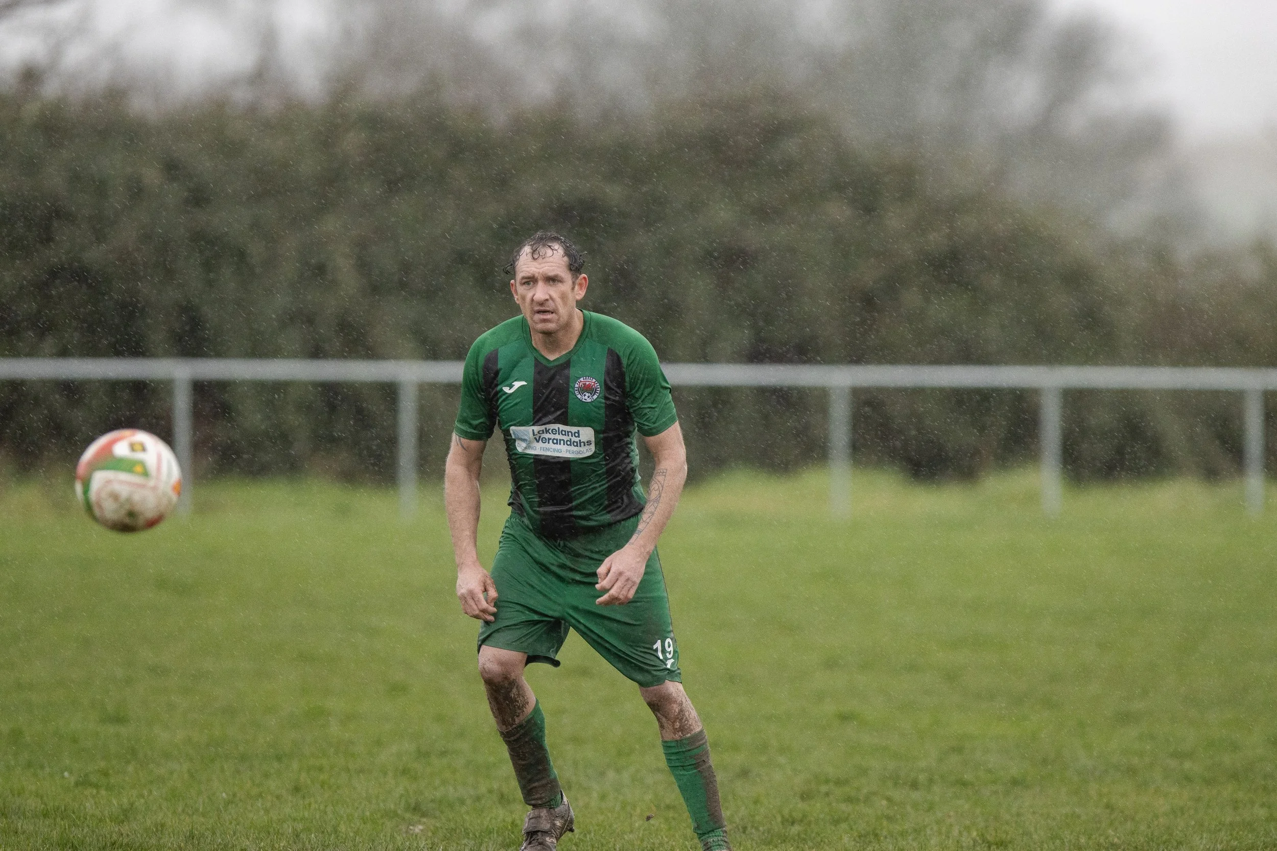 A soccer player in a green and black uniform is on a grassy field in the rain, with a soccer ball in the air to the left, and a blurred background of trees and a fence.