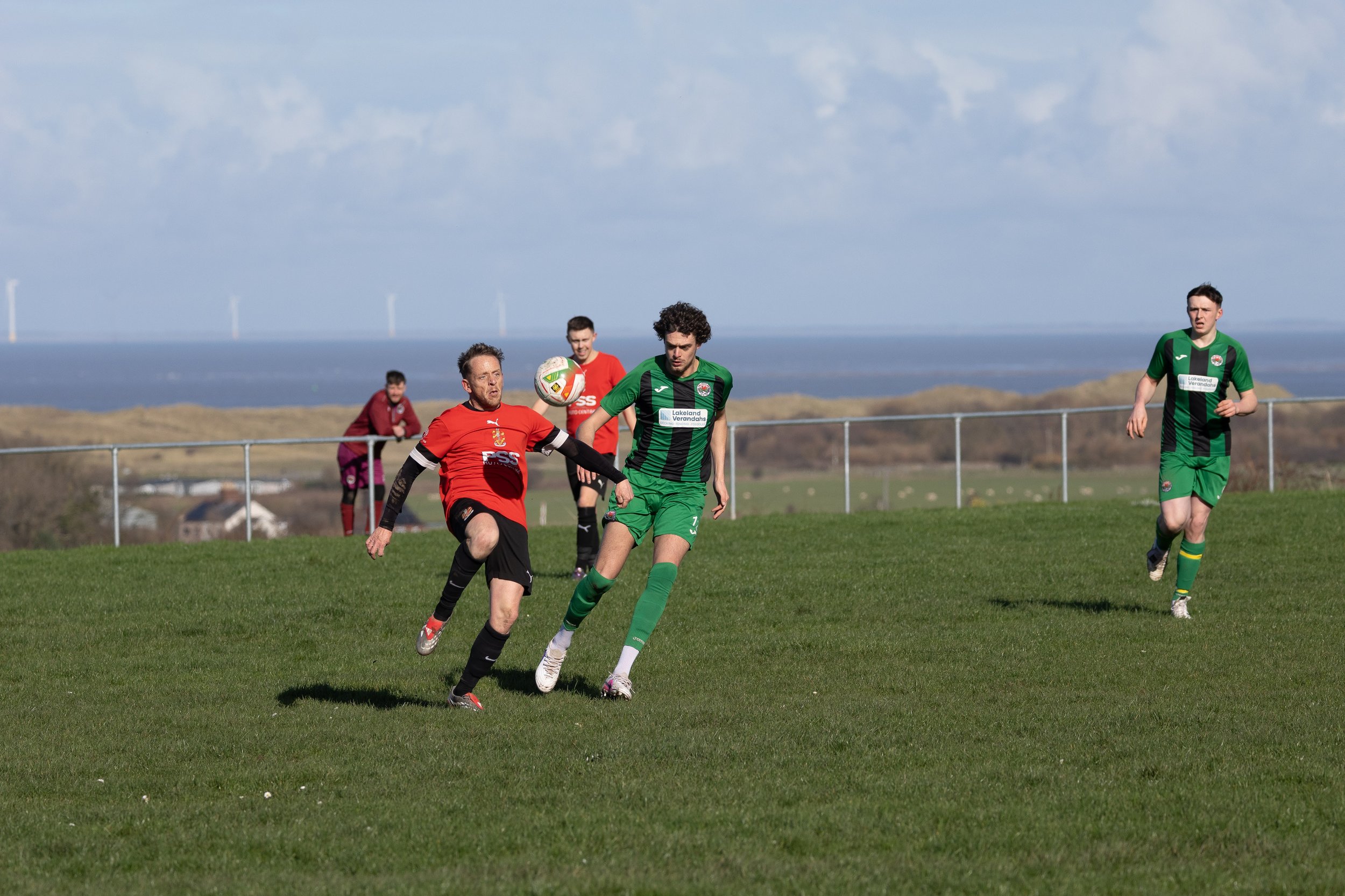 Soccer players competing for the ball on a grassy field with wind turbines and a cloudy sky in the background.
