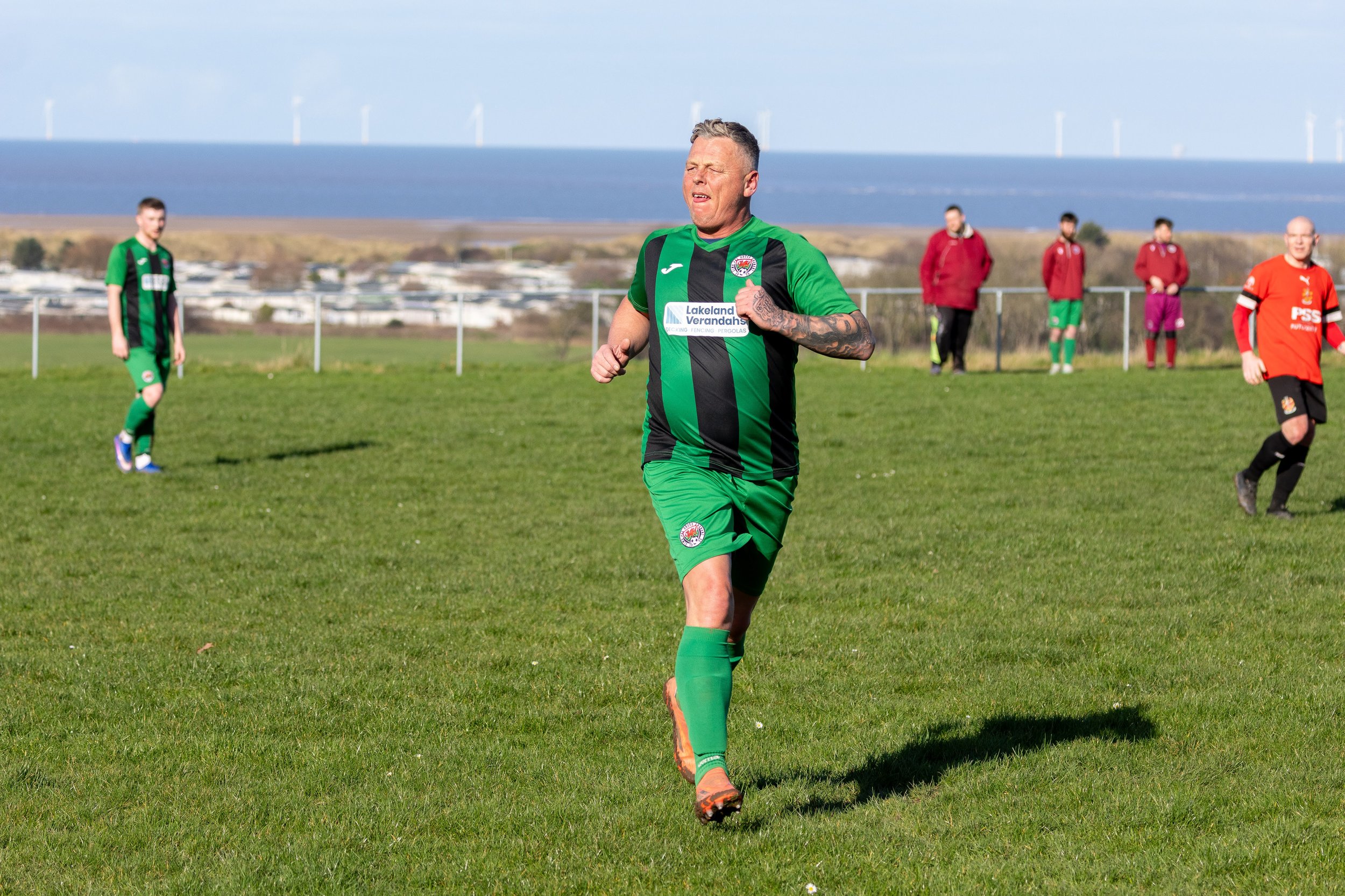 A man in a green and black soccer uniform running on a grass field, with four other players and three spectators in the background, and wind turbines in the distance.