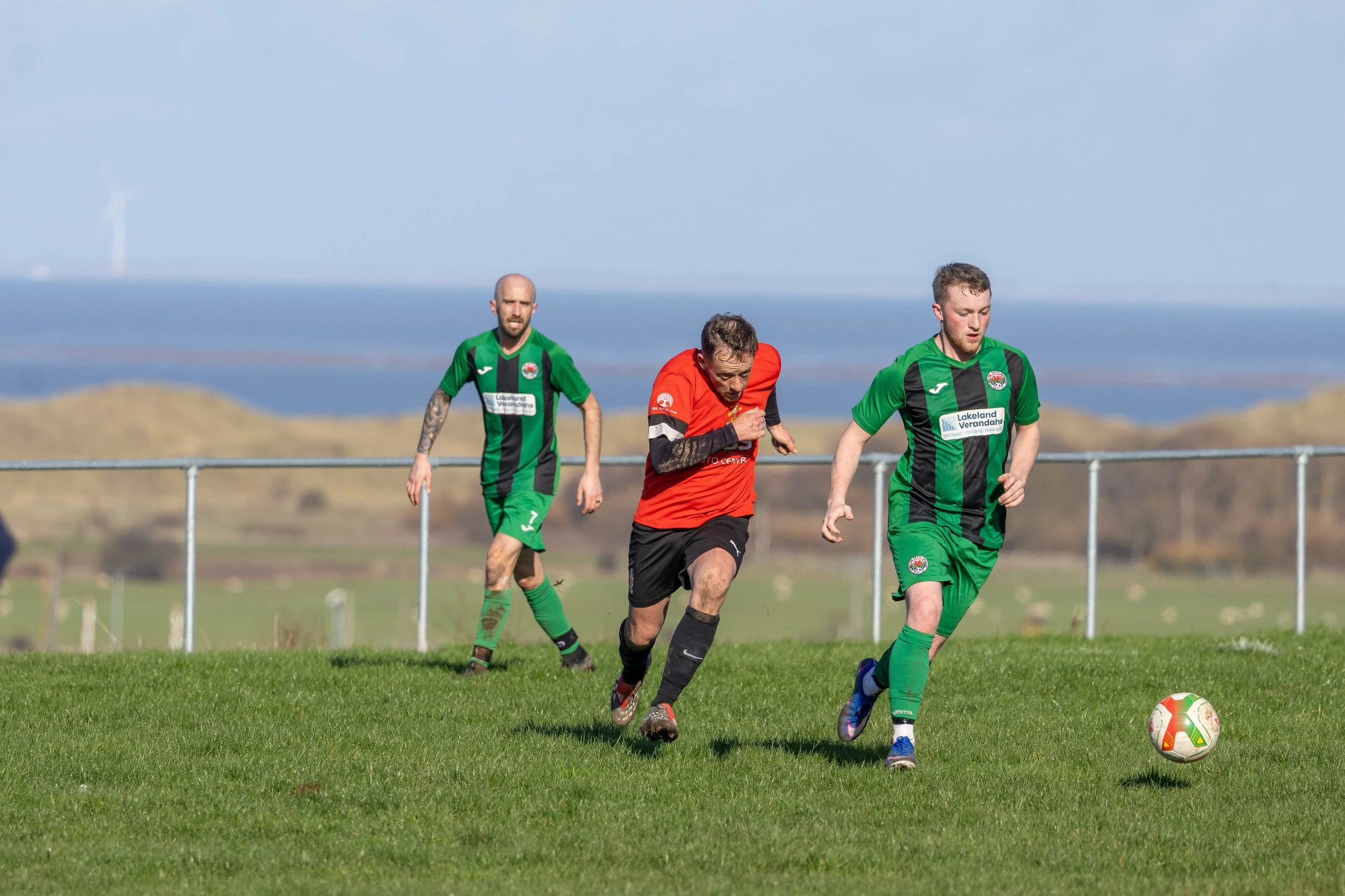 Three men playing soccer on a grassy field with a scenic landscape in the background.