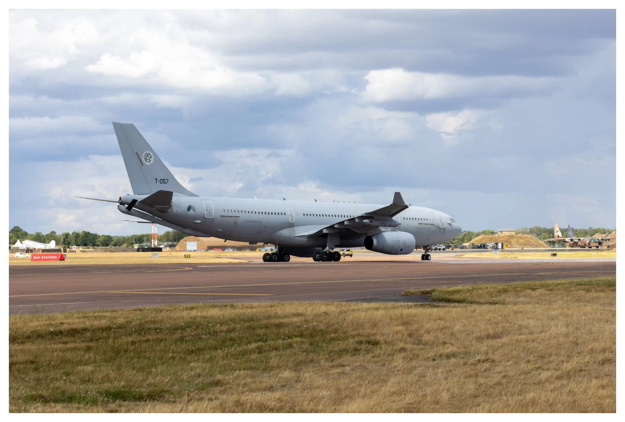 A large military cargo airplane on the runway at an airport, with a cloudy sky overhead and other aircraft and airport structures in the background.
