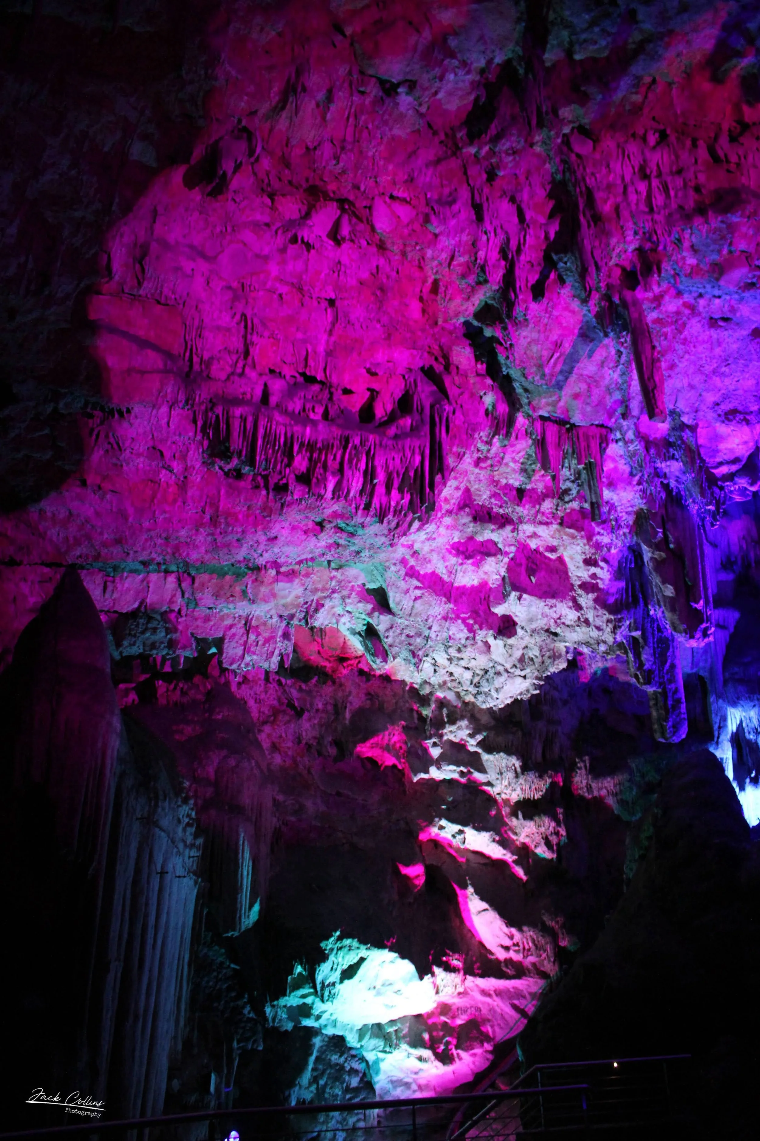Colorfully lit cave with pink, purple, and blue lights illuminating stalactites and stalagmites.