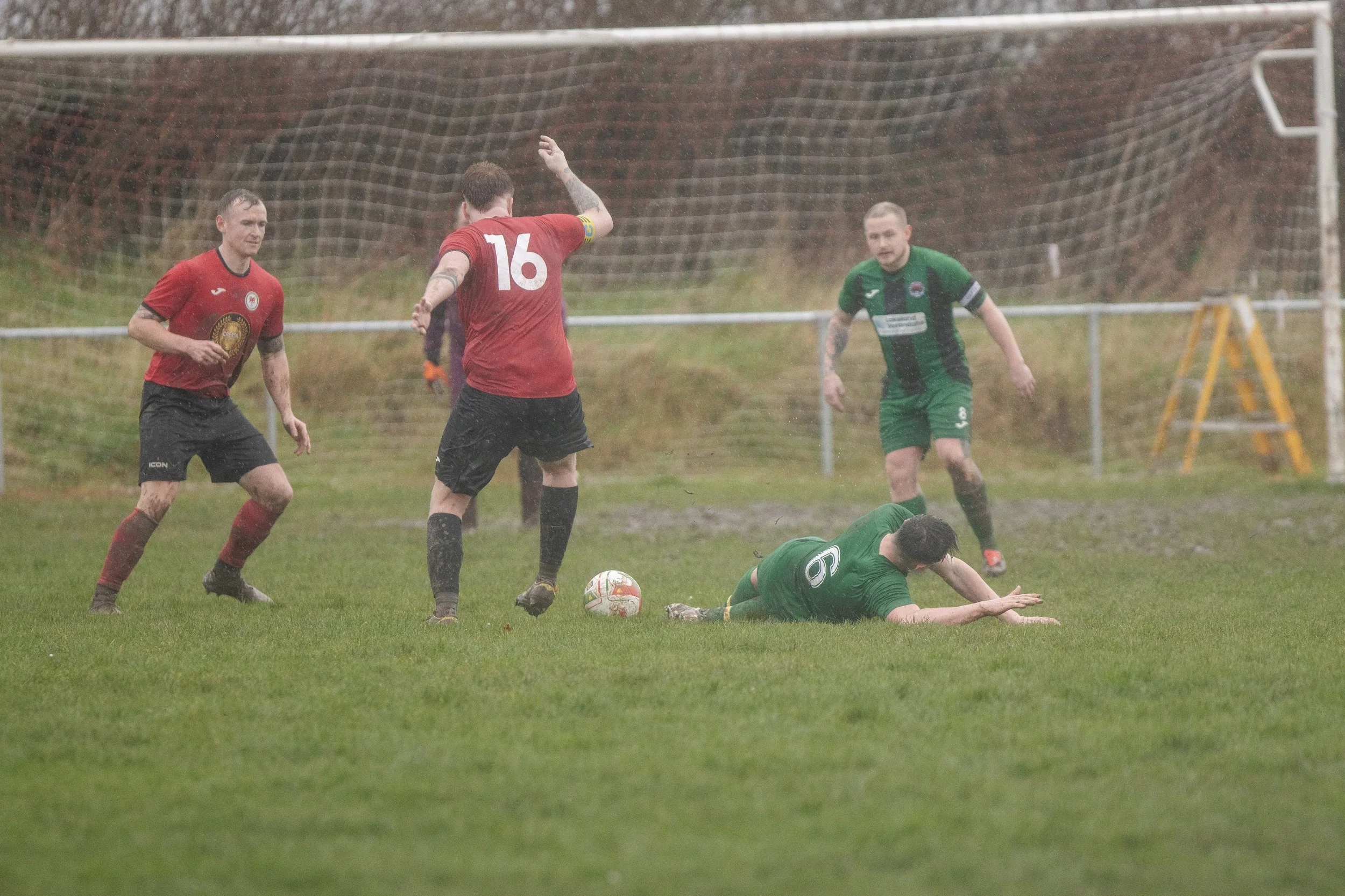 A rainy soccer match showing players from two teams, one in green and the other in red, with a player in green lying on the ground near the goal as others move nearby.
