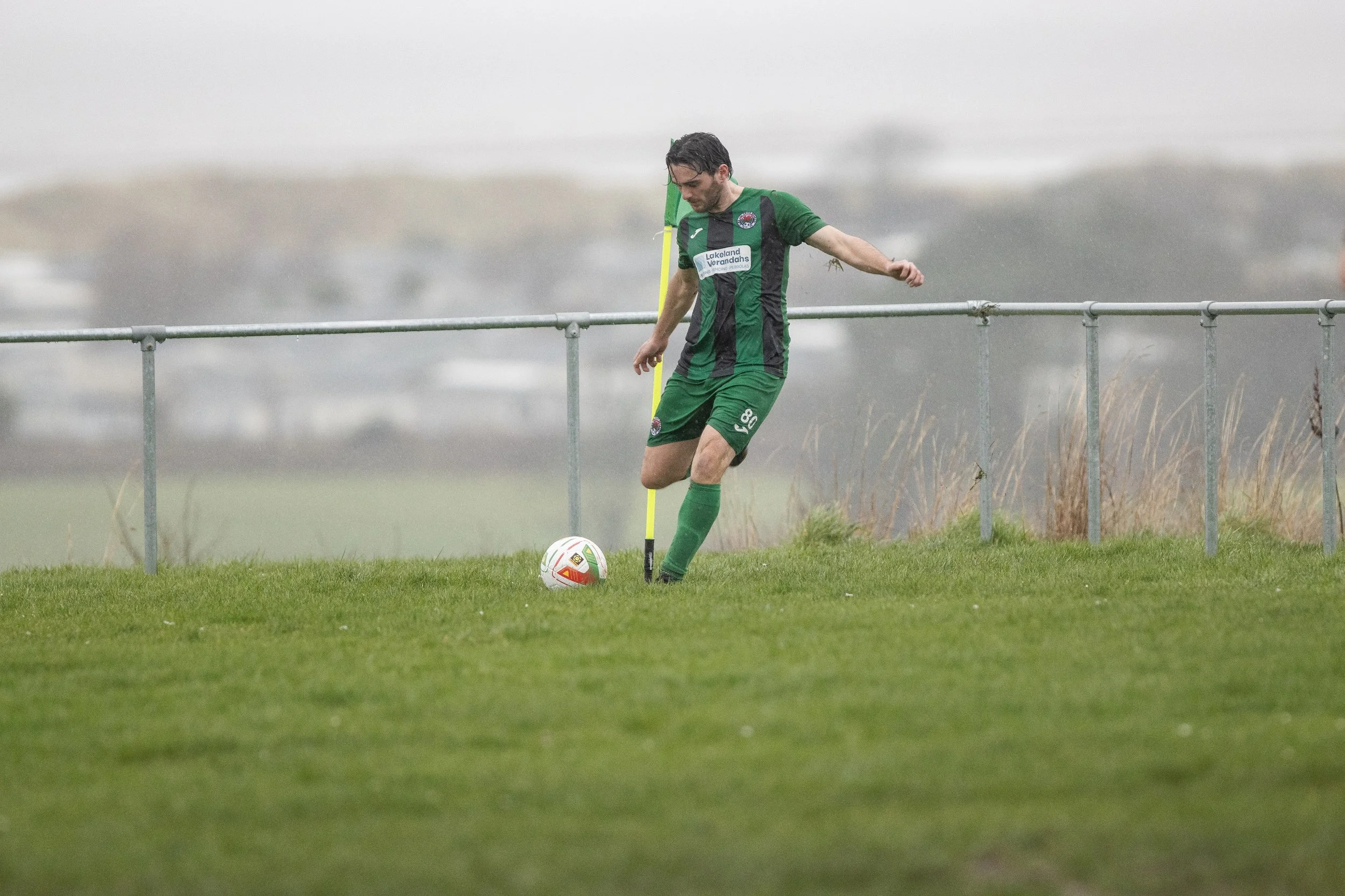 A soccer player in green and black uniform practicing on a grassy field on a rainy day.