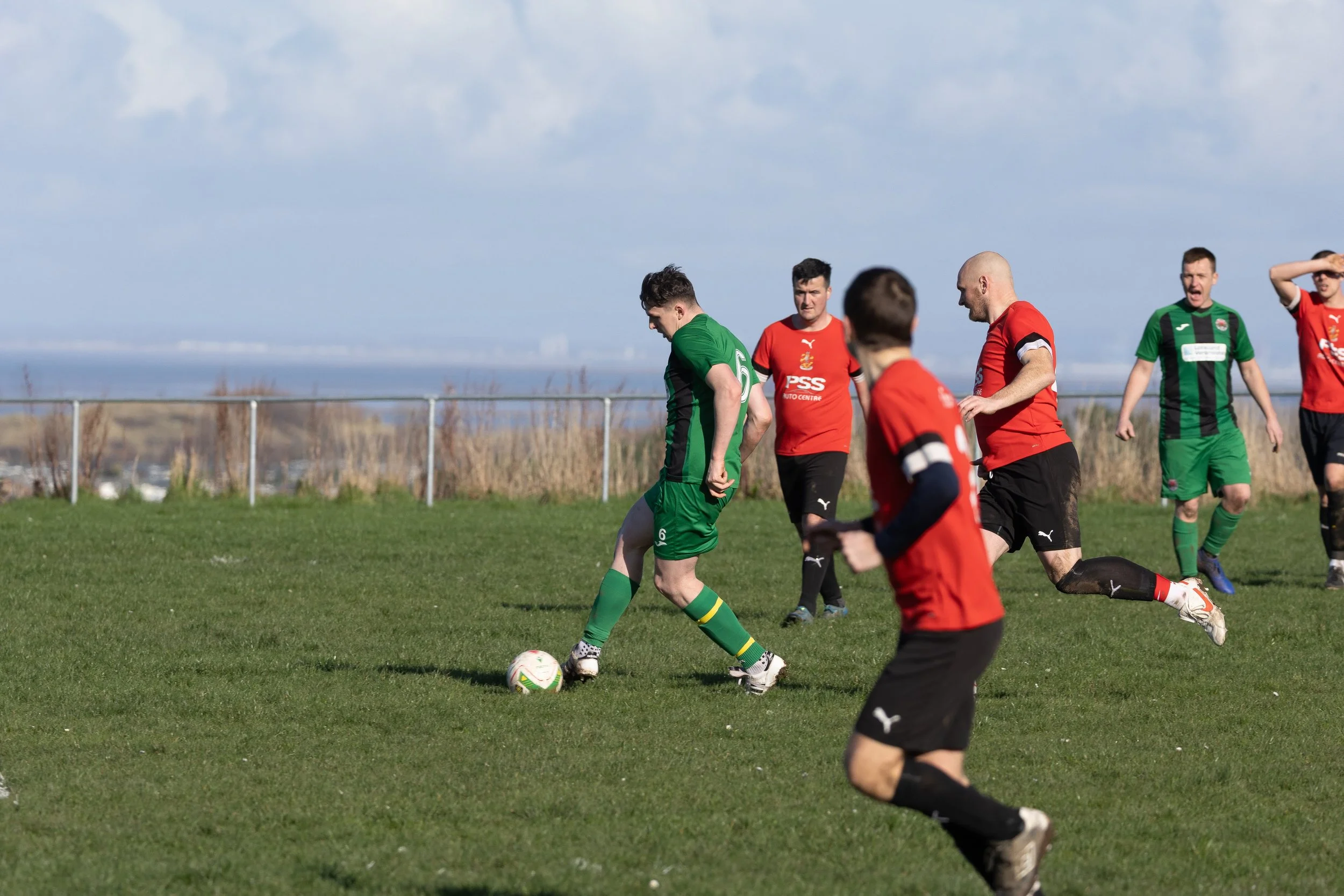 Soccer players on a grassy field during a match, with some players in red jerseys and others in green jerseys, under a partly cloudy sky, with a metal fence and landscape in the background.
