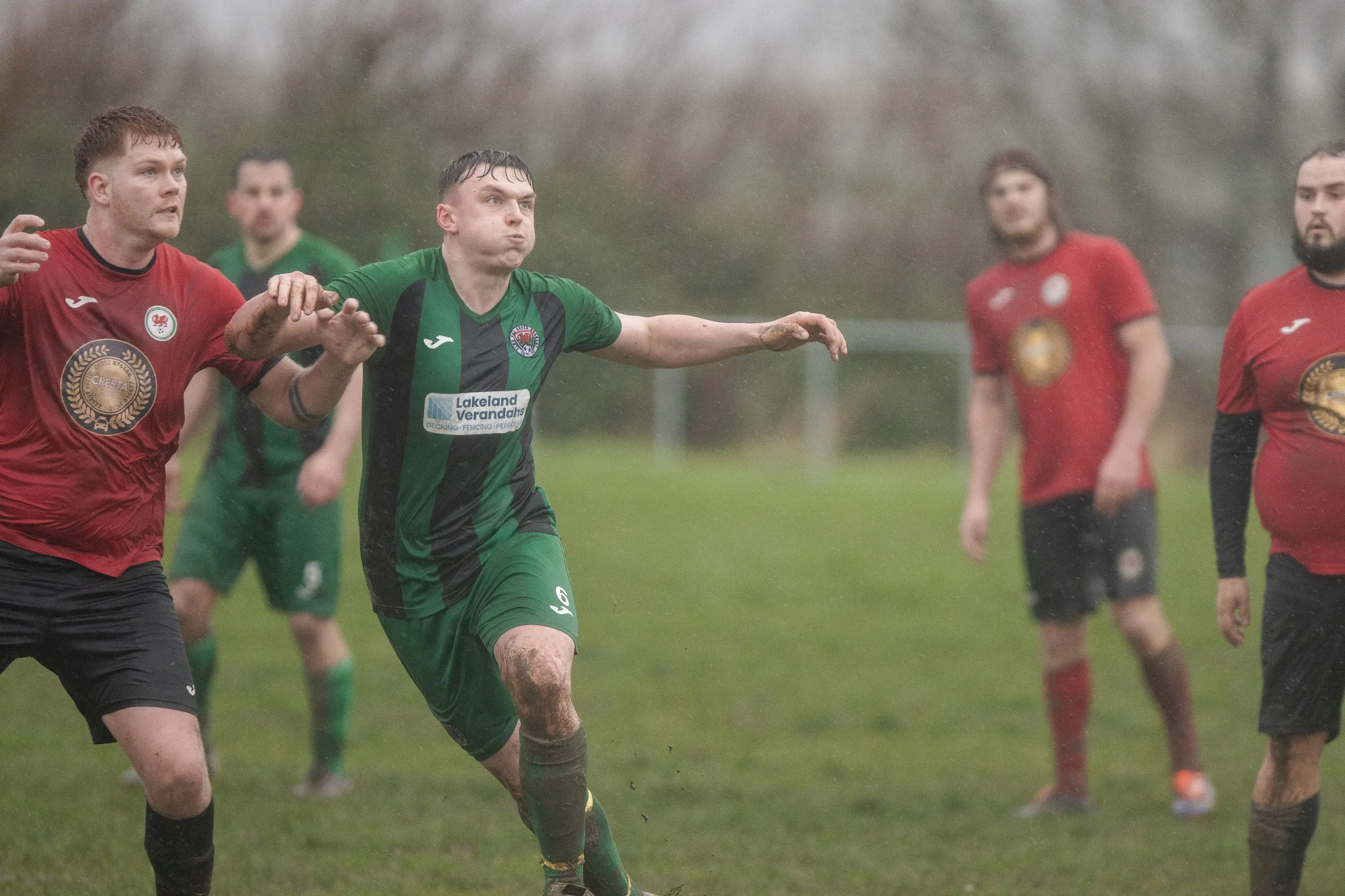 Soccer players competing on a muddy field in rainy weather, with some players wearing red jerseys and one in green and black, all appearing dirty and wet.