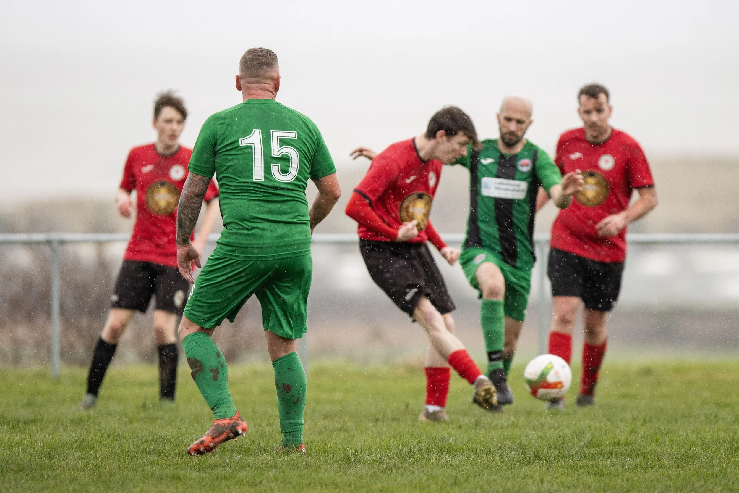 Soccer players play against each other on a muddy field in rainy weather. The players are wearing red and green uniforms, with muddy shoes and wet grass visible.