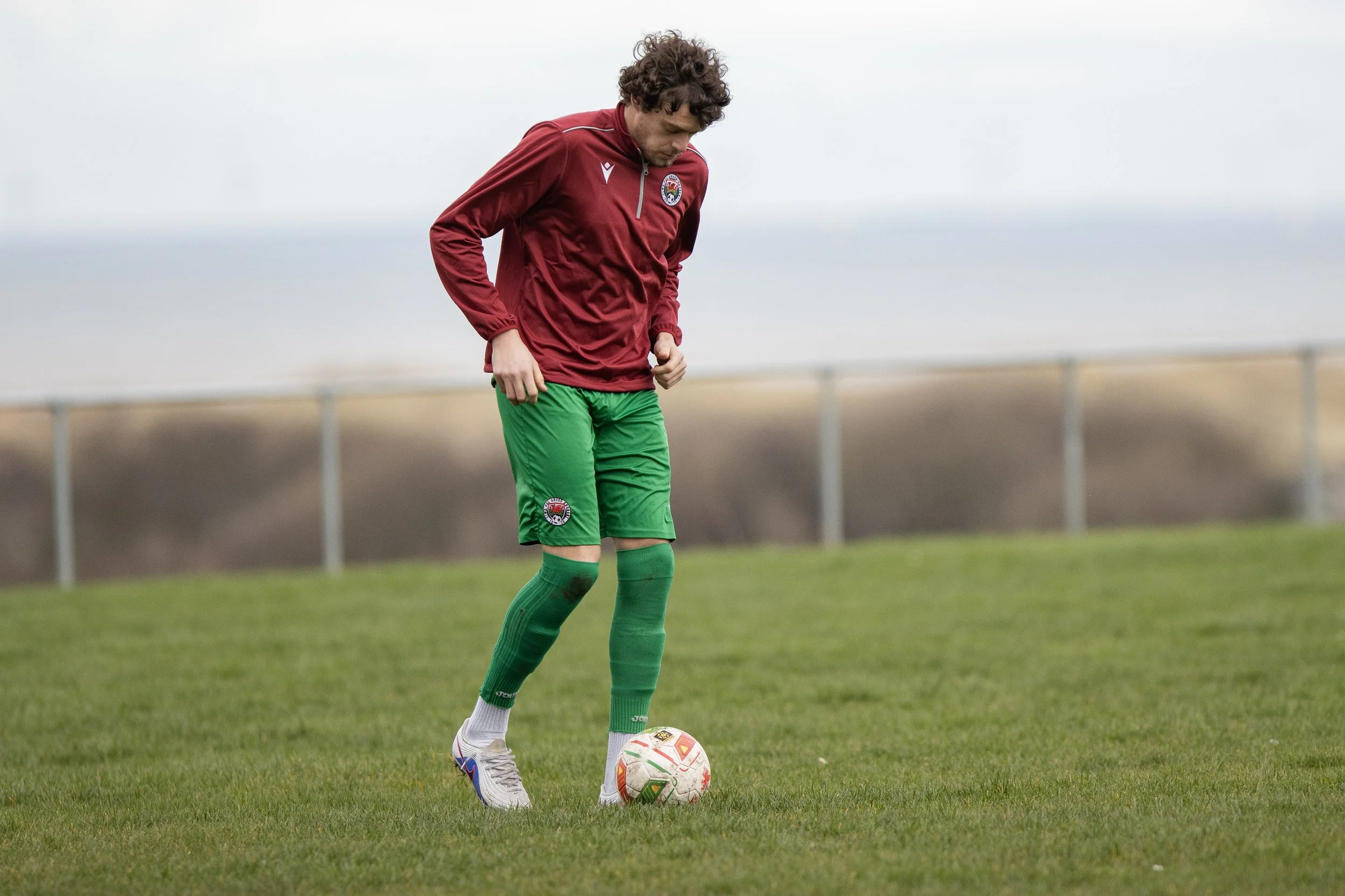 A soccer player wearing a maroon jacket and green shorts standing on a grass field, looking down at a soccer ball.