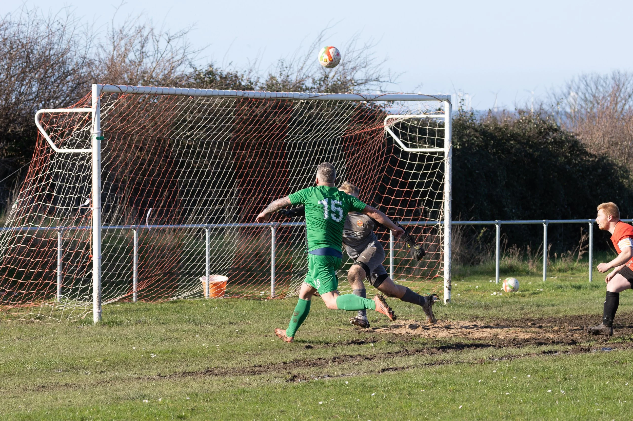 Soccer match with players near the goal, one in a green jersey with number 15, and a goalkeeper in black, with a ball in mid-air heading towards the goal.