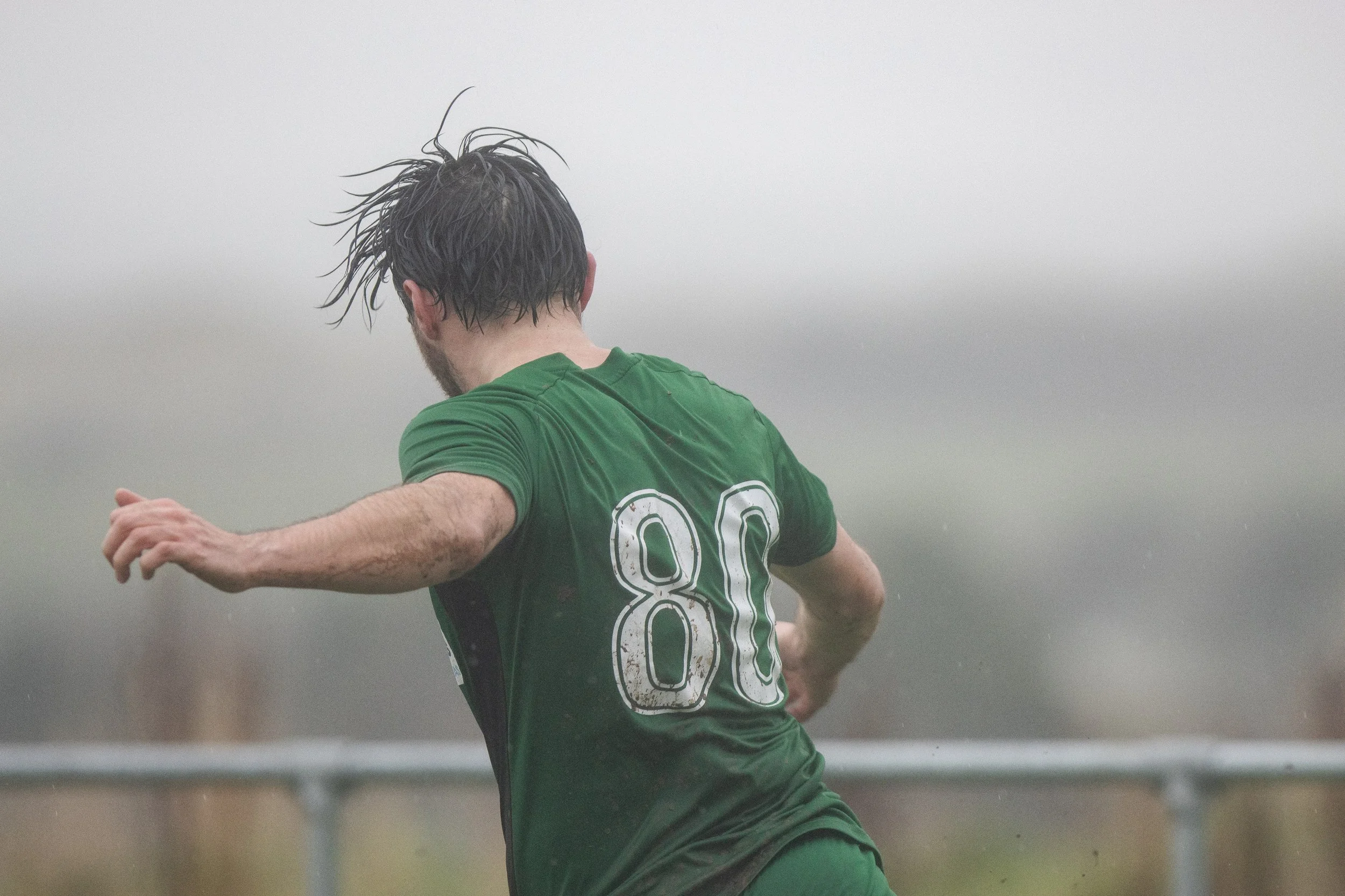 A man wearing a green soccer jersey with the number 80 on the back, running outdoors in the rain with wet hair and muddy arms, facing away from the camera.