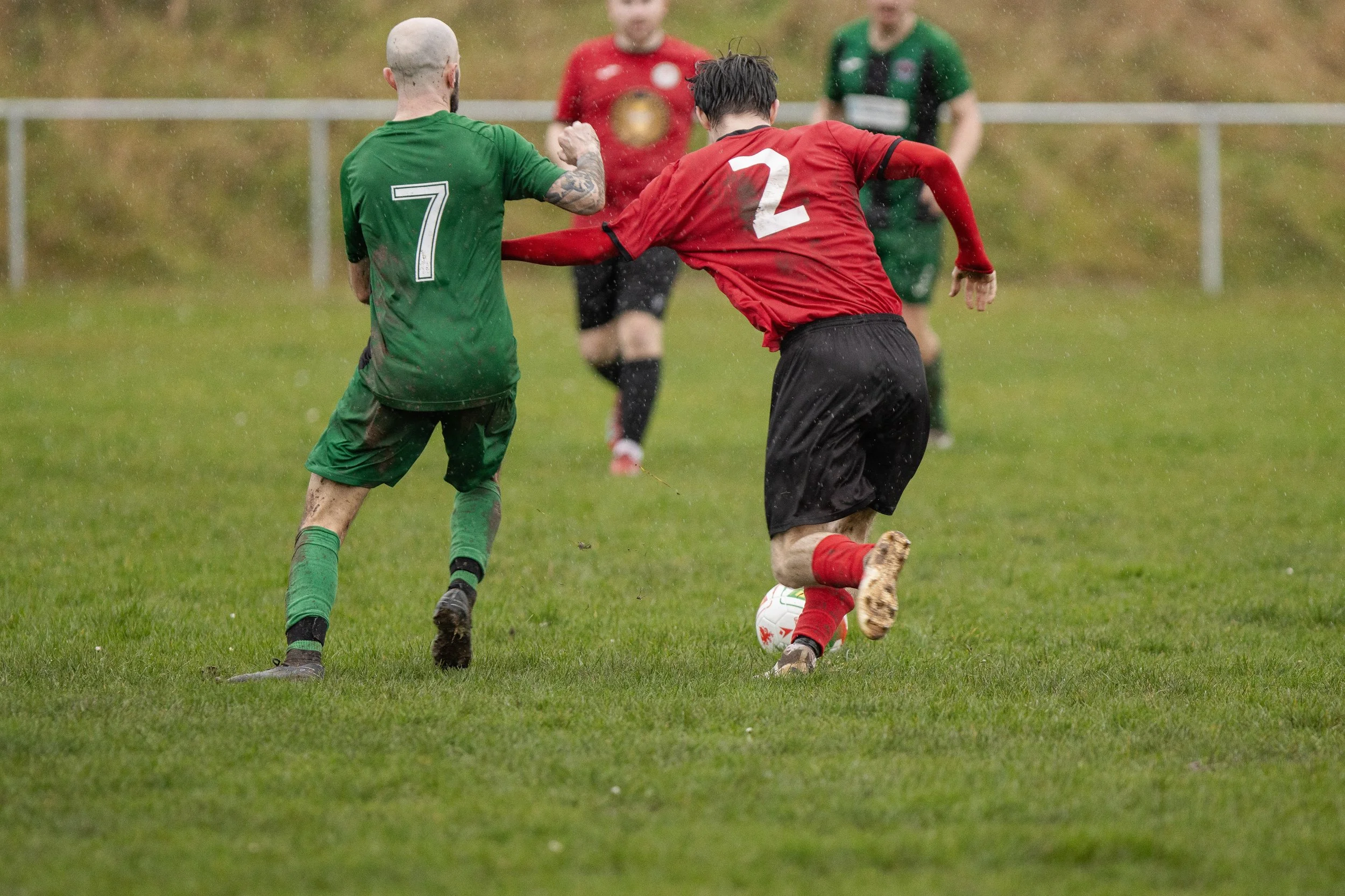 Two soccer players, one in a green uniform with the number 7 and the other in a red uniform with the number 2, compete for the ball on a wet grass field. Two other players are visible in the background, one in a red uniform and the other in a green u