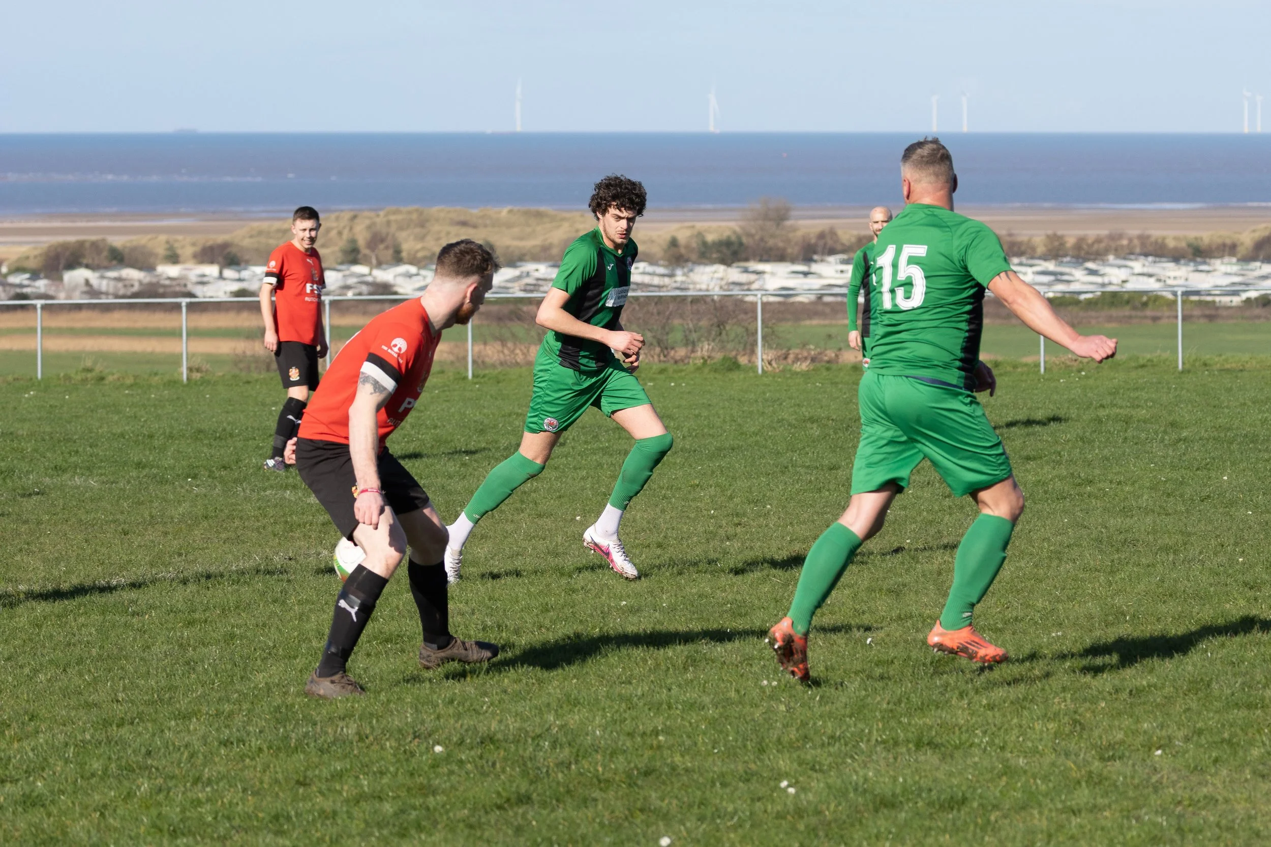 Soccer players on a grassy field near the coast, with ocean and wind turbines in the background. Players are wearing green and red uniforms.