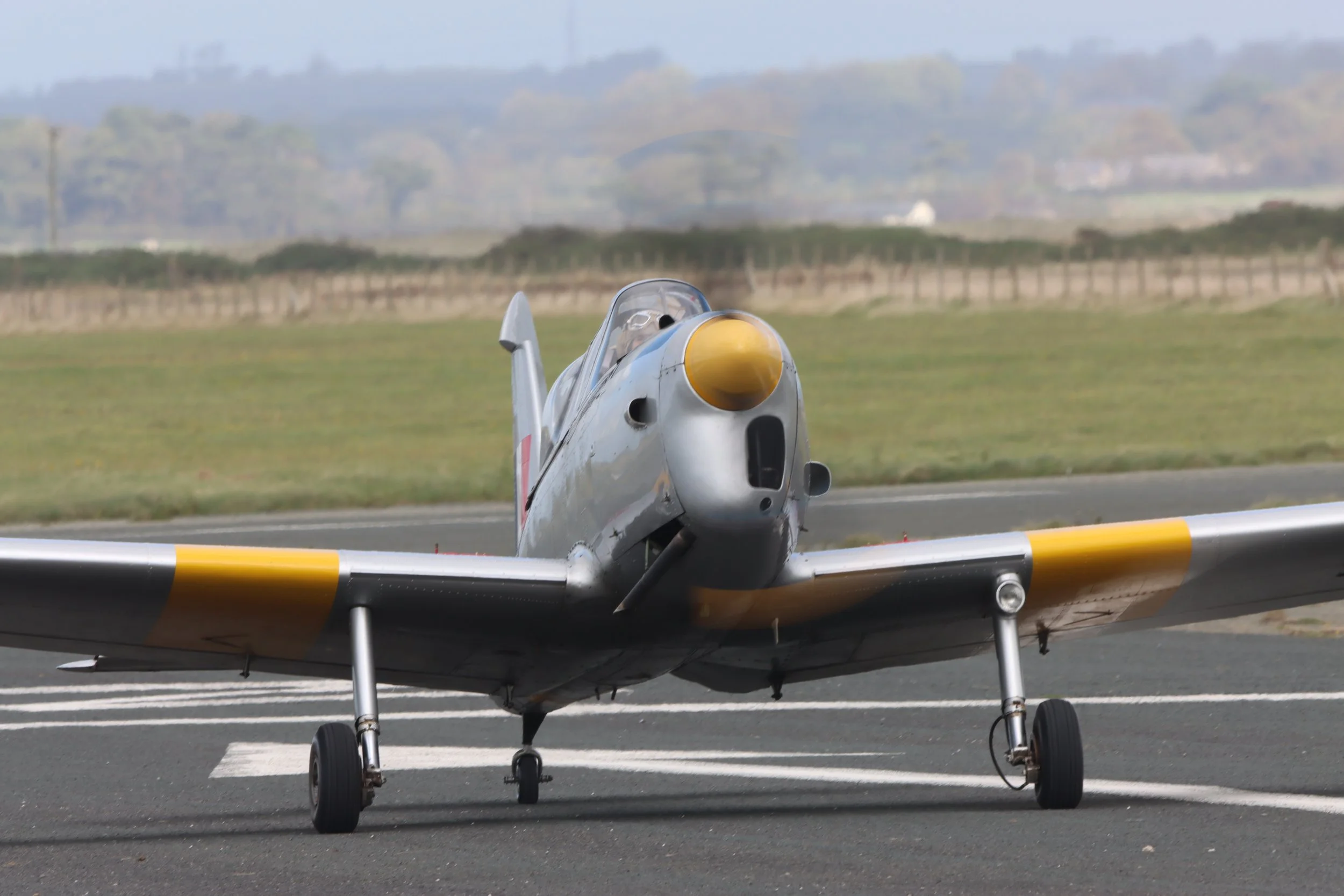 A vintage jet aircraft on a runway with a yellow nose cone and landing gear, with grassy fields and a distant tree-lined landscape in the background.