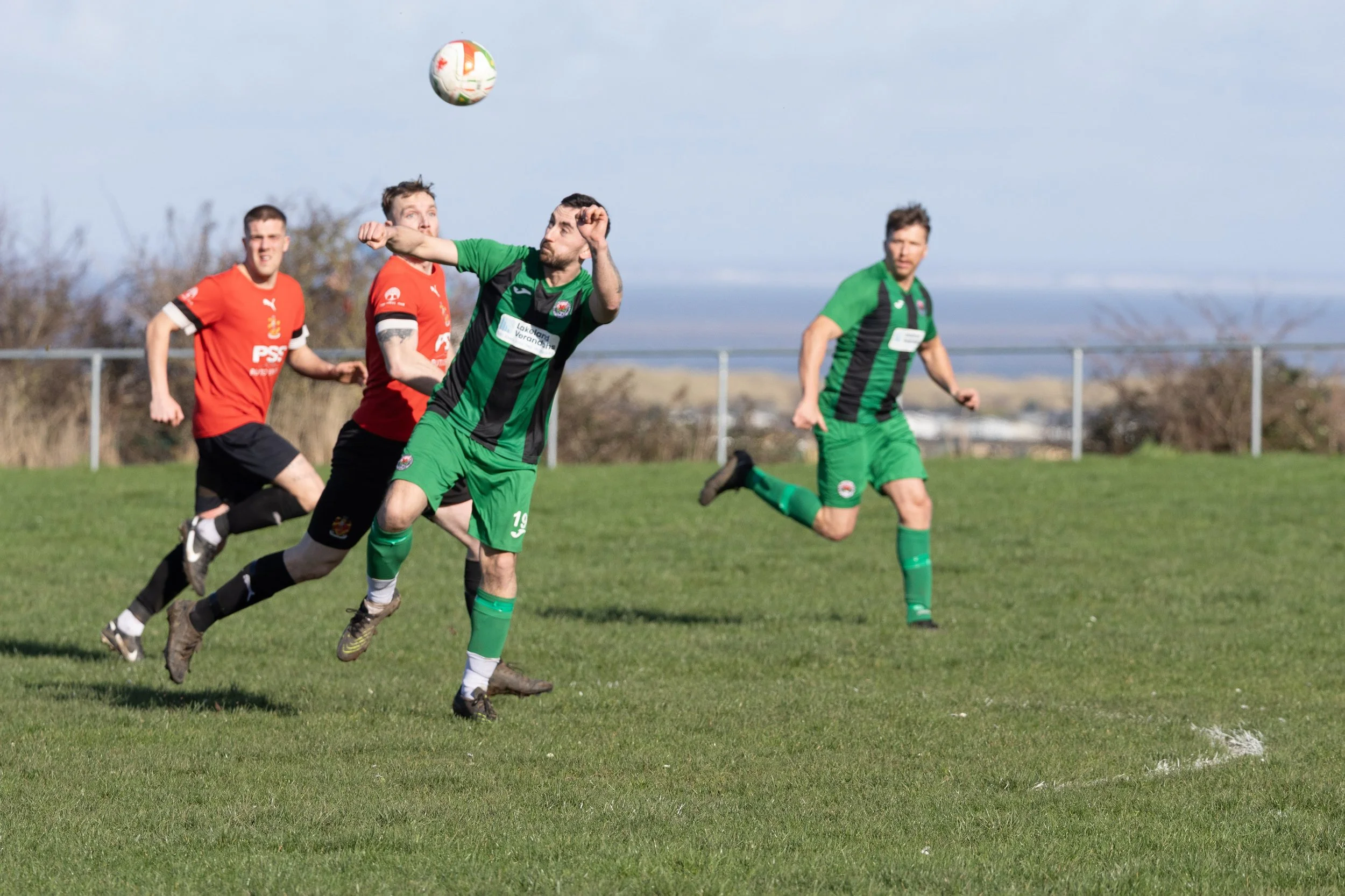 Four men playing soccer on a grass field, two wearing red jerseys and two wearing green jerseys with black stripes, with a soccer ball in the air.