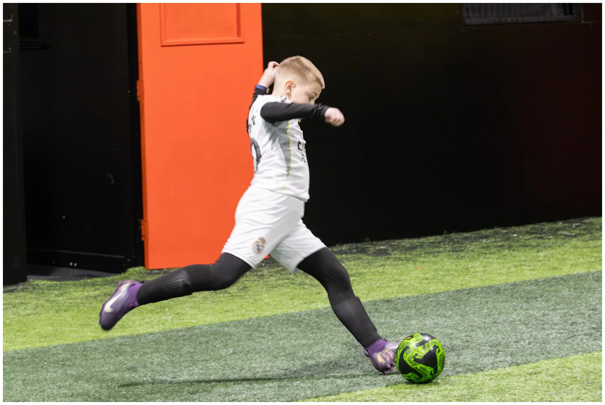 Young boy in a soccer uniform kicking a green and black soccer ball on an indoor field.