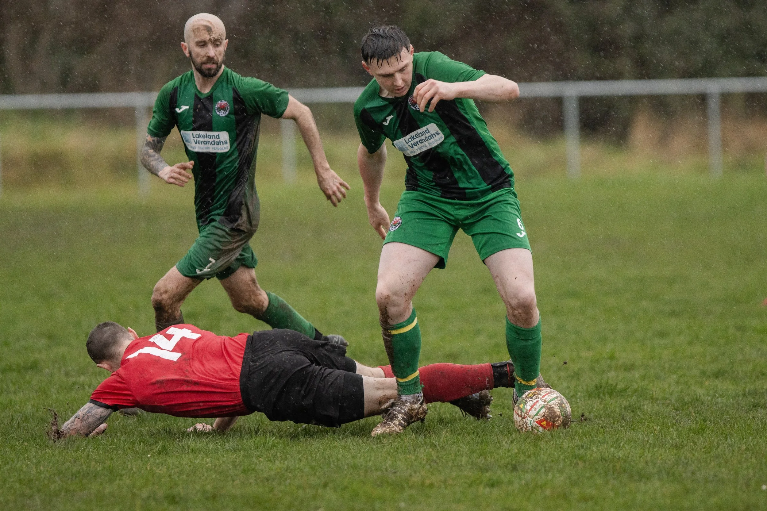 Two soccer players in green jerseys compete for the ball while a player in red and black dives on the ground during a match on a grassy field, with mud and rain visible.