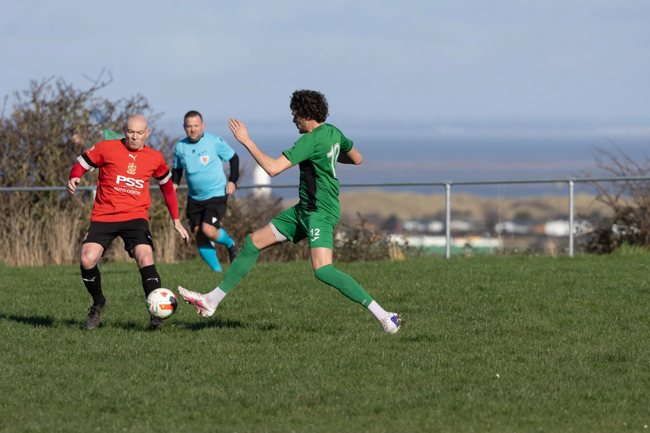 Soccer players in a match, one in a red jersey and one in a green jersey, competing for the ball on a grassy field with a fence and distant landscape in the background.