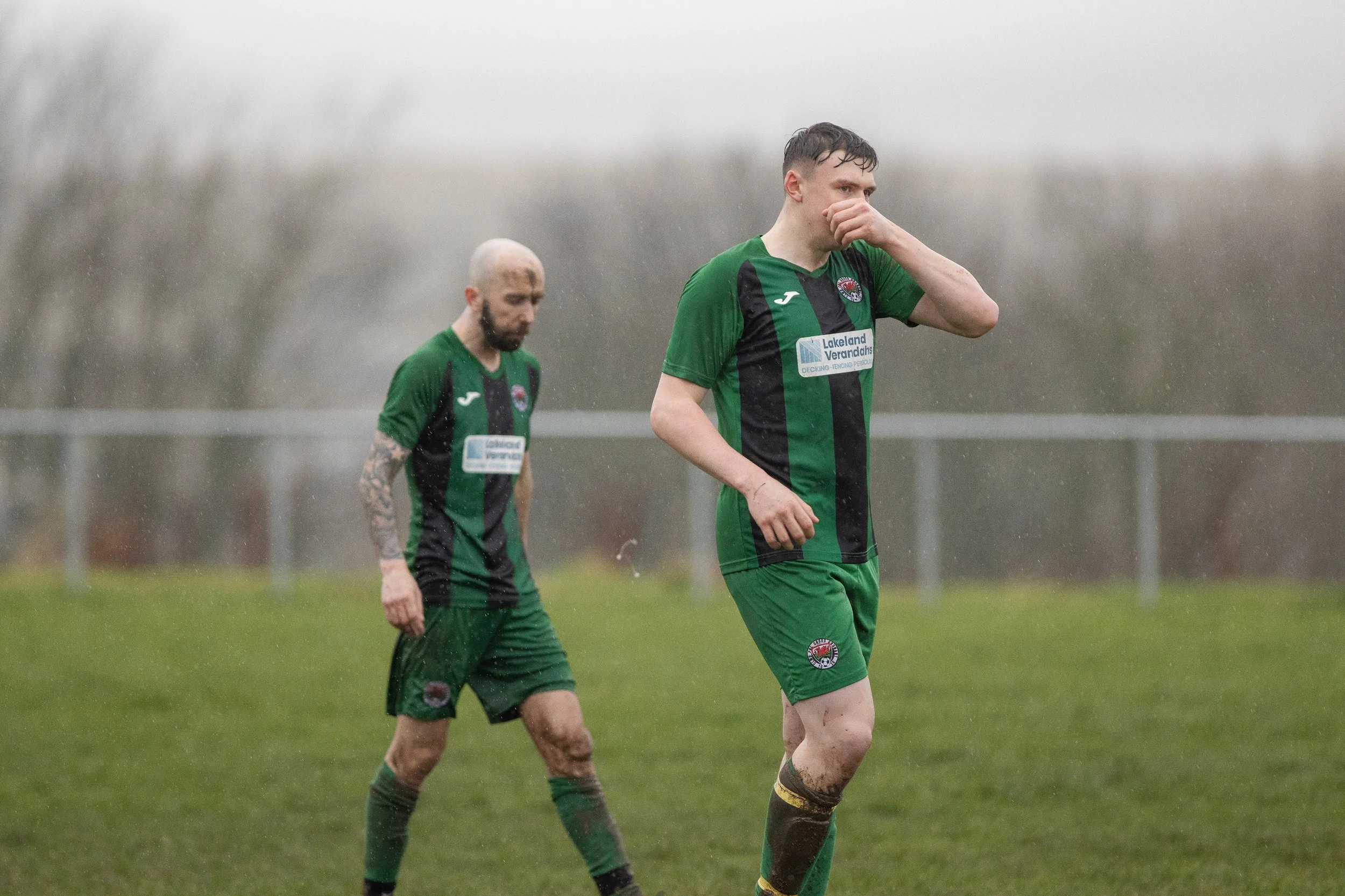 Two soccer players walking on a rainy field wearing green and black uniforms, with one covering his face with his hand.