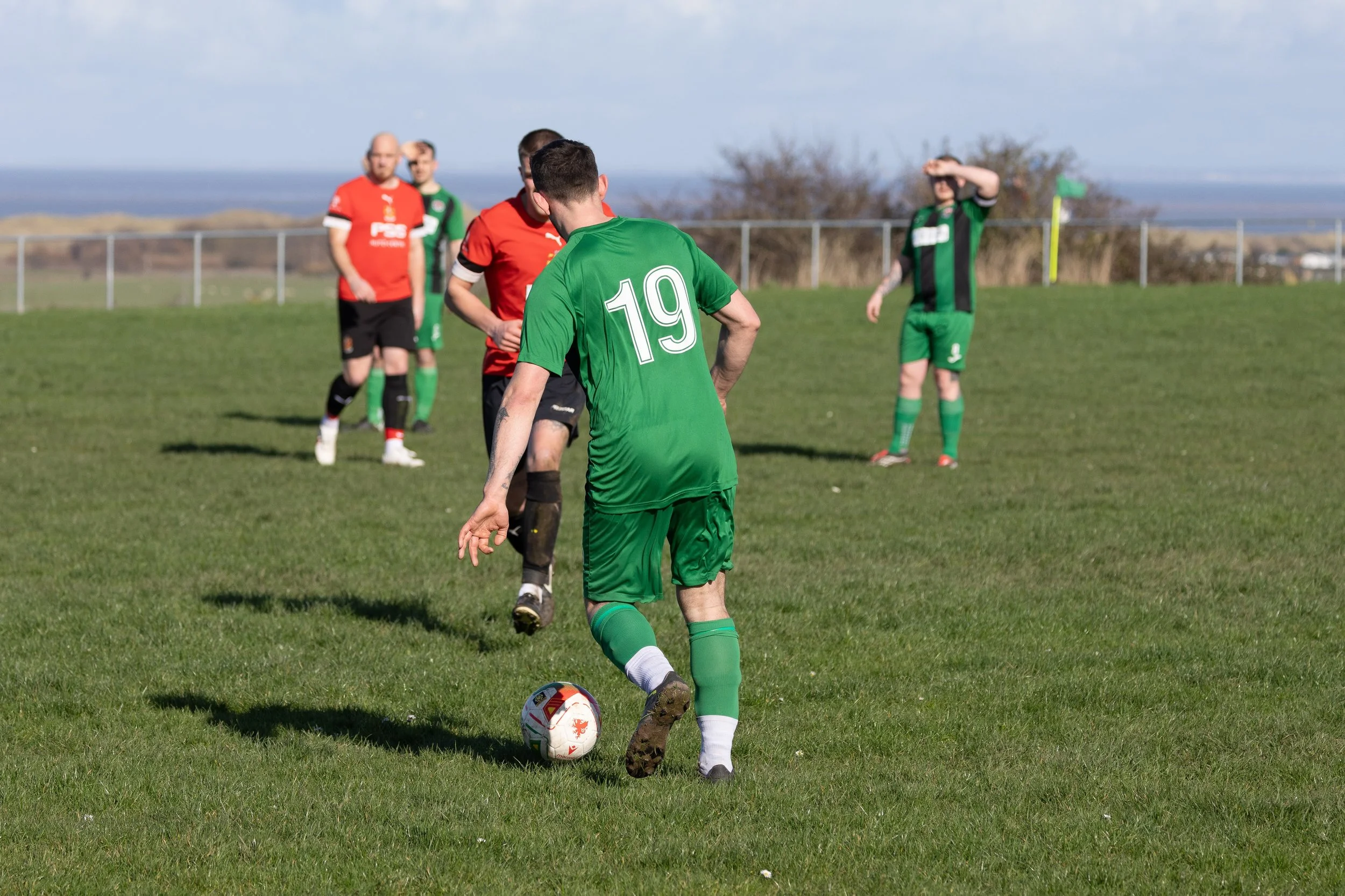 Soccer players on a grassy field, with one player in a green uniform about to kick the ball, and other players in red and green in the background.