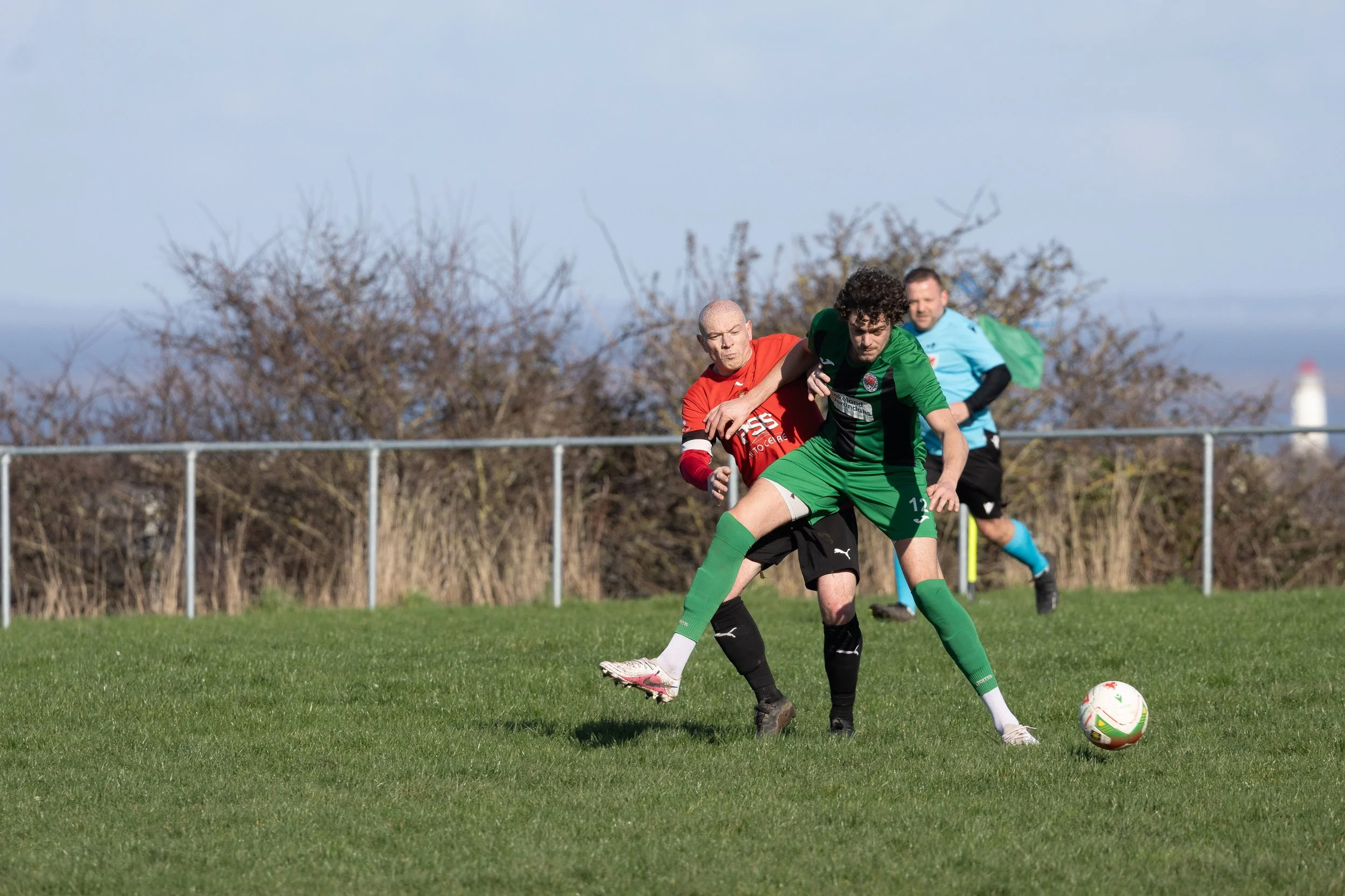 Soccer players competing for the ball during a match on a grassy field, with a fence and trees in the background.