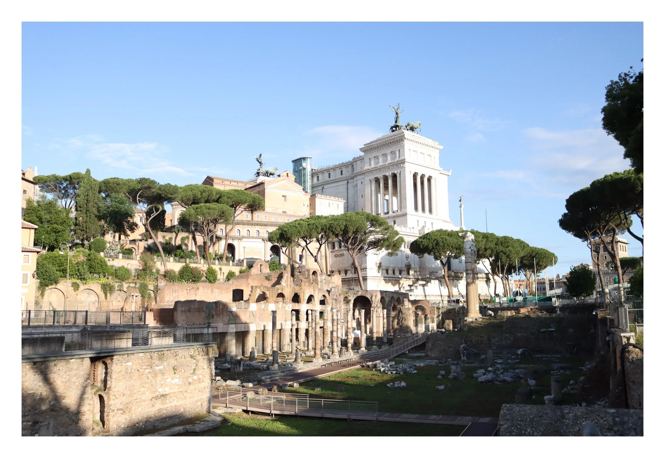 Ancient Roman ruins with columns and stone structures in the foreground, trees on a hill, and the Altare della Patria monument in Rome, Italy, in the background. Clear blue sky.