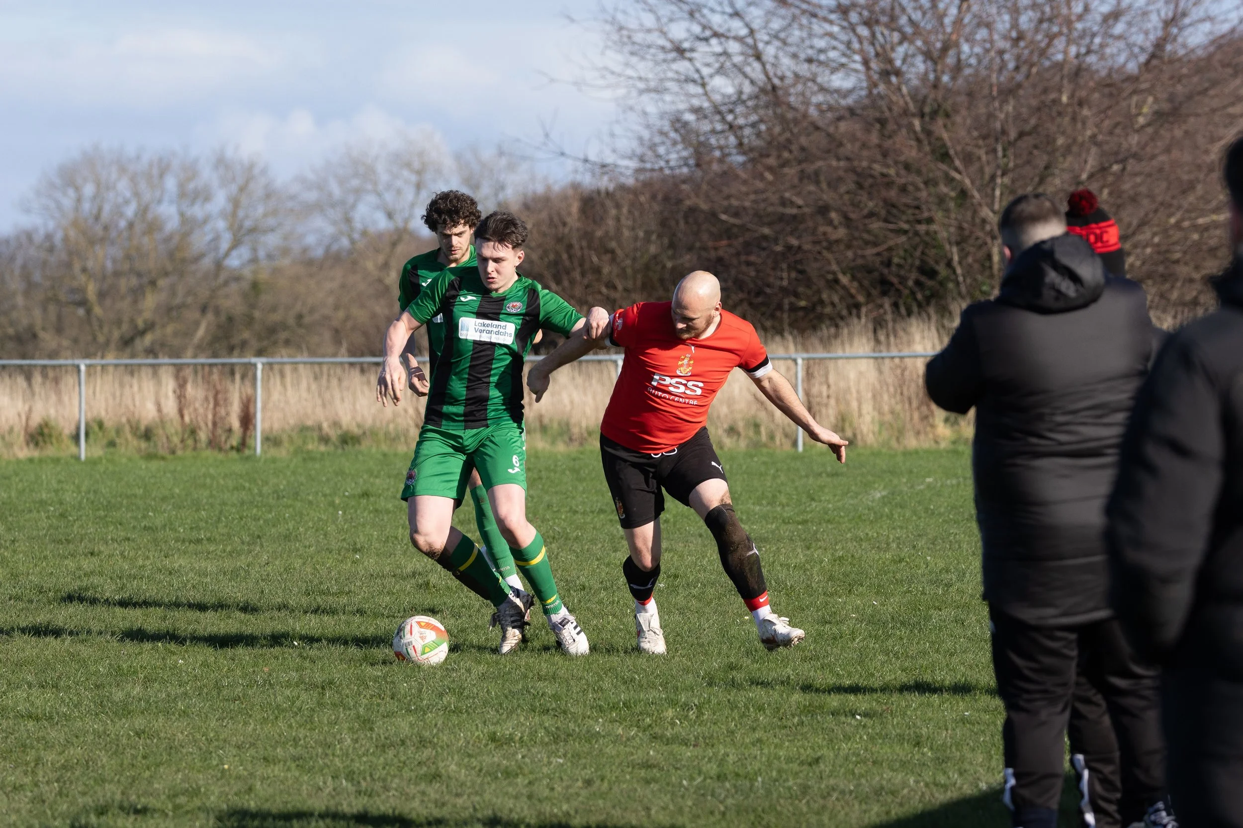 Two soccer players contest for the ball on a grassy field during a match, with spectators watching from the sidelines.