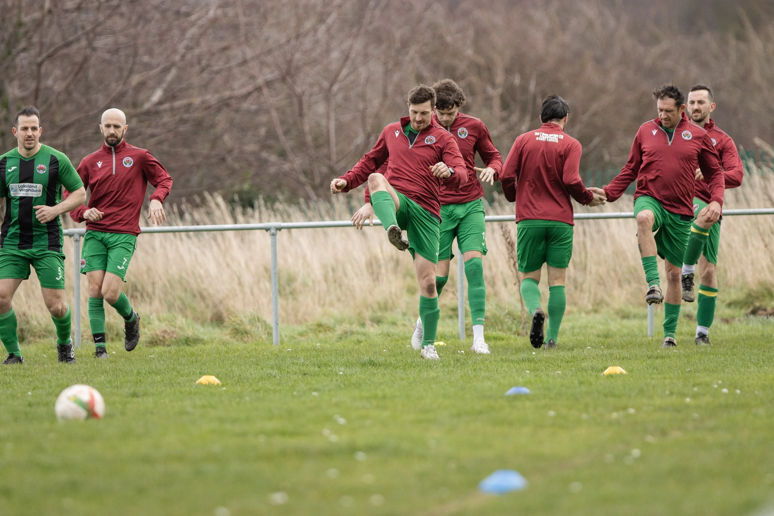 Soccer players warming up on a grassy field, some stretching and jogging, with a marker ball and cone in the foreground, and trees in the background.