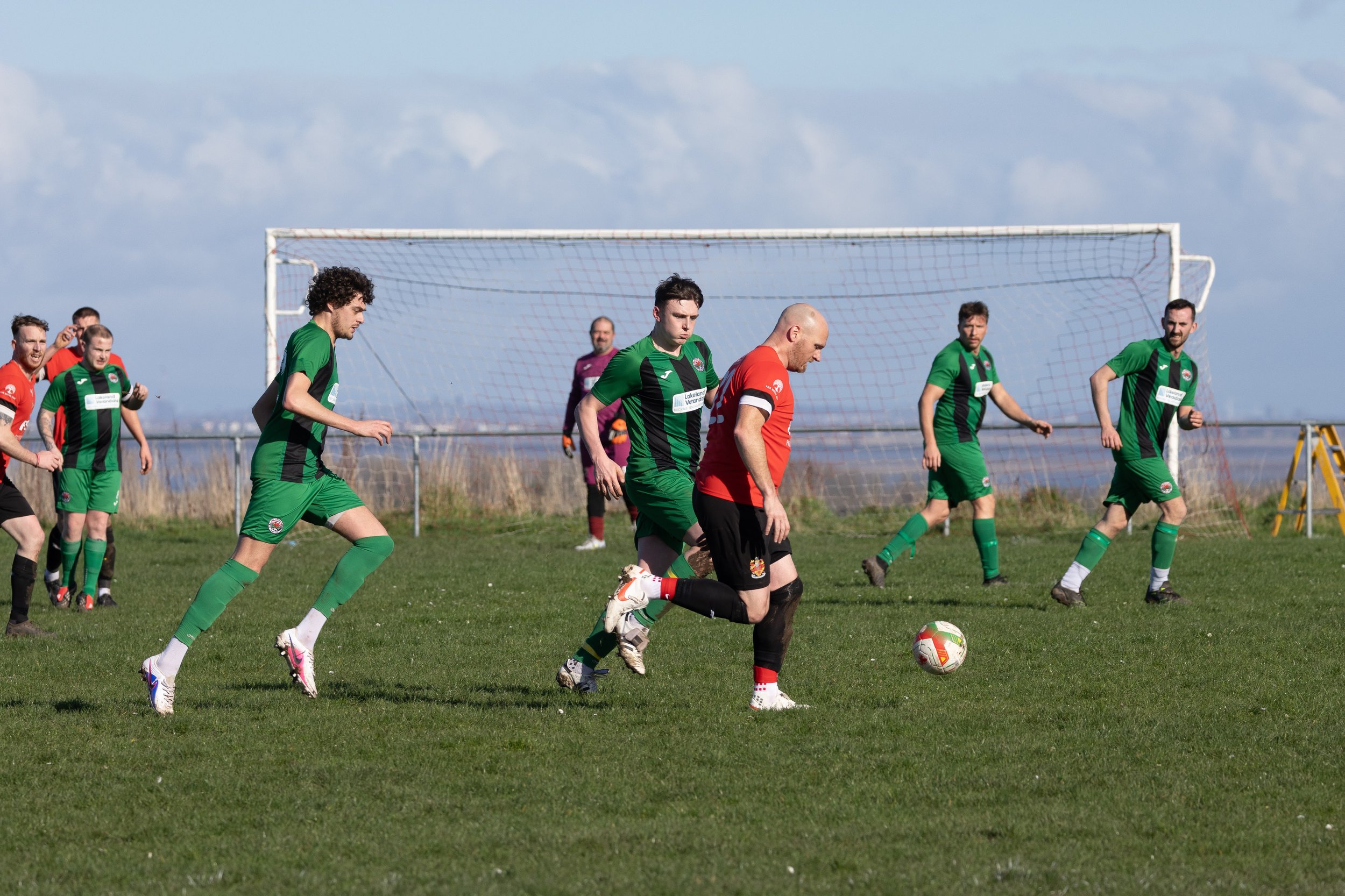 Soccer match with players in green and black jerseys and an opponent in red, on a grassy field near a goal, under a partly cloudy sky.