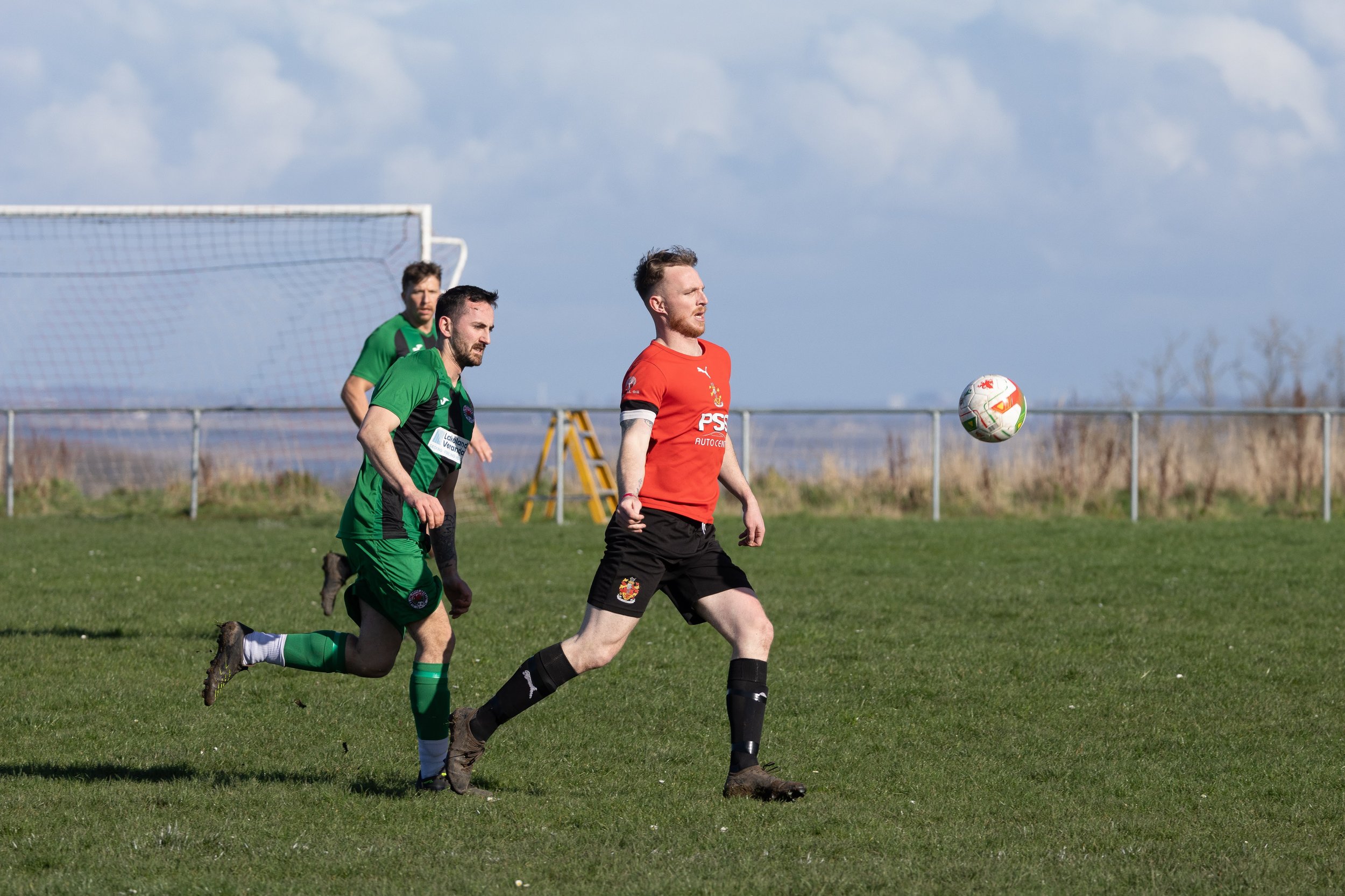 Three soccer players on a grassy field during a game, with one in a red jersey and two in green jerseys, and a soccer ball in front of the player in red.
