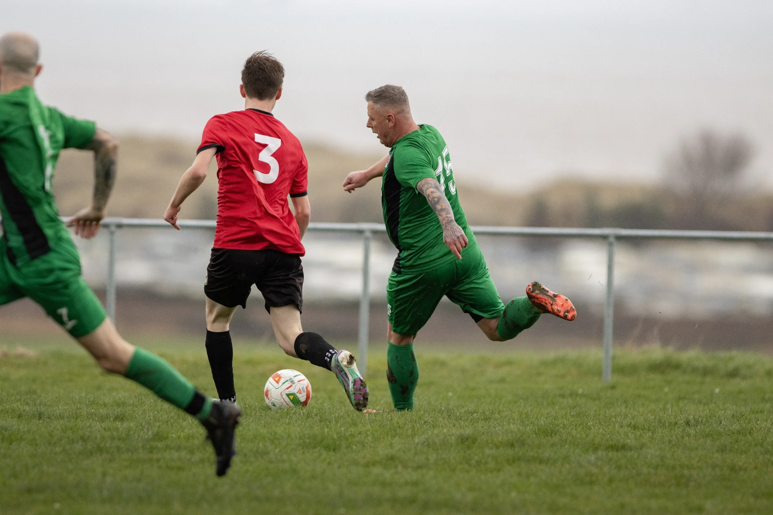 Soccer players in green and red jerseys competing for the ball on a grassy field with a metal fence in the background.