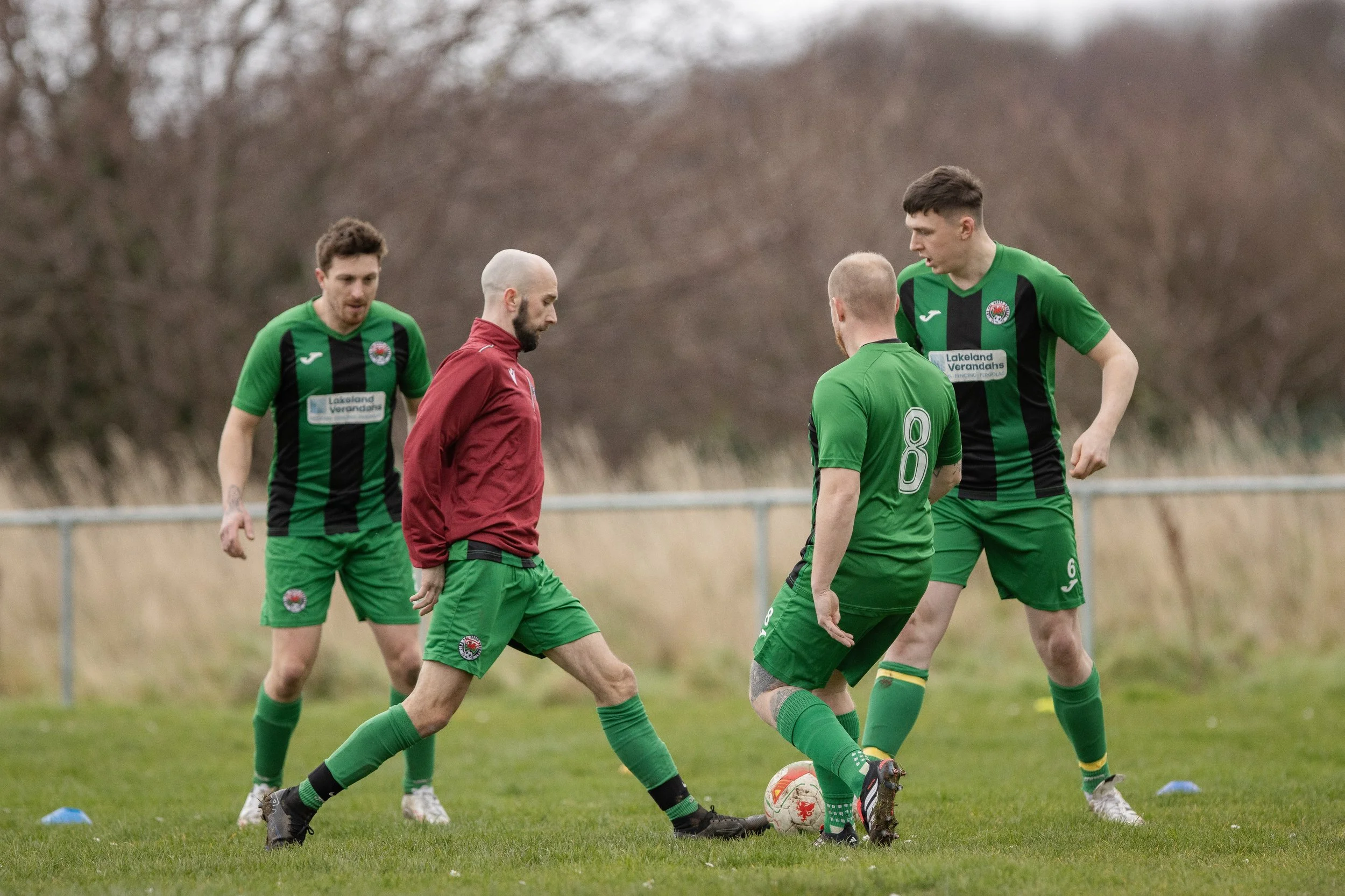 Four men playing soccer on a grass field, two wearing green jerseys with black stripes, one in a red jacket, and one in green shorts with a number 8 on the back, standing around a soccer ball.