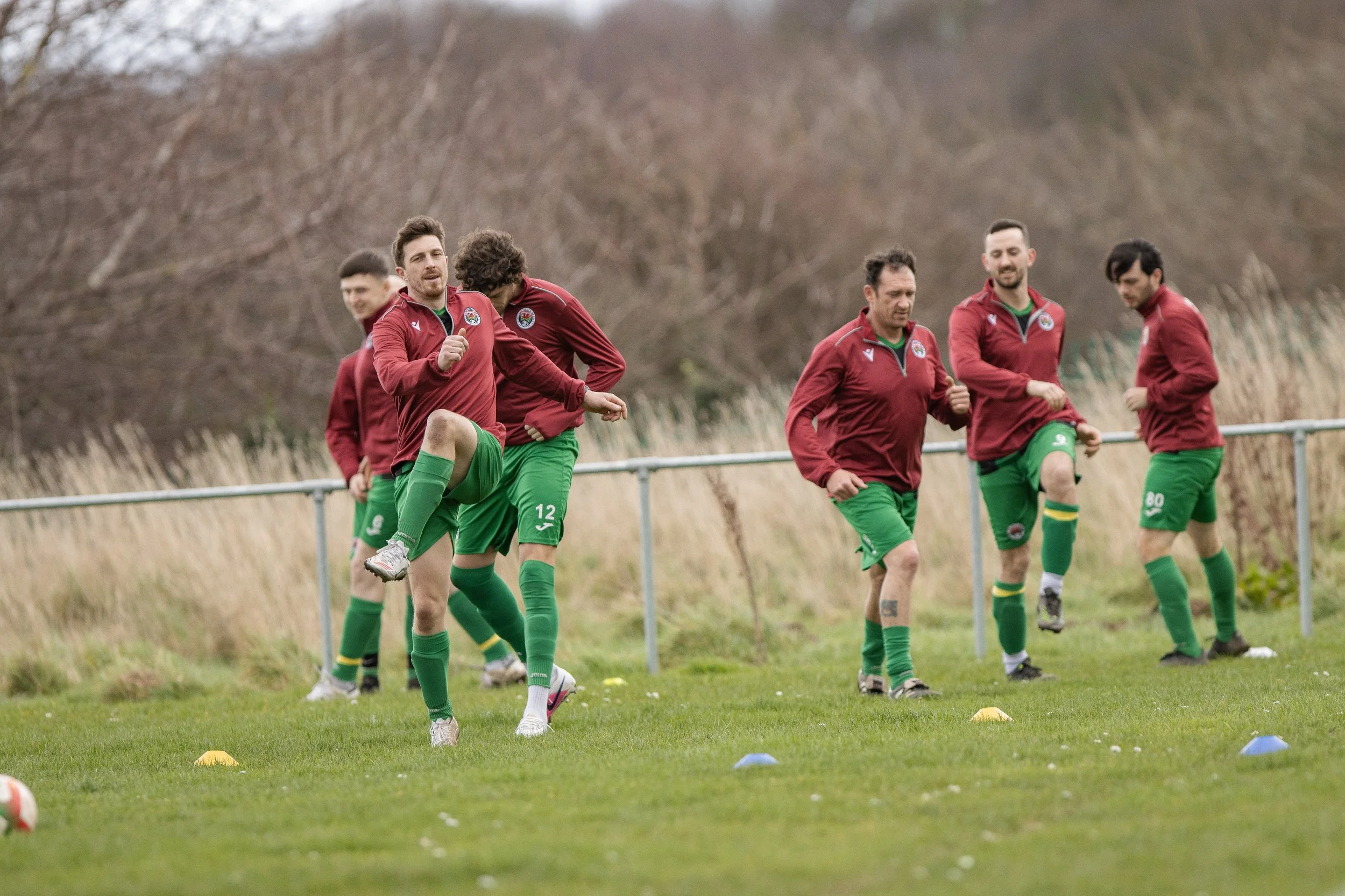 A group of six men dressed in athletic gear warming up on a grassy field with a metal fence and leafless trees in the background, suggesting outdoor training.